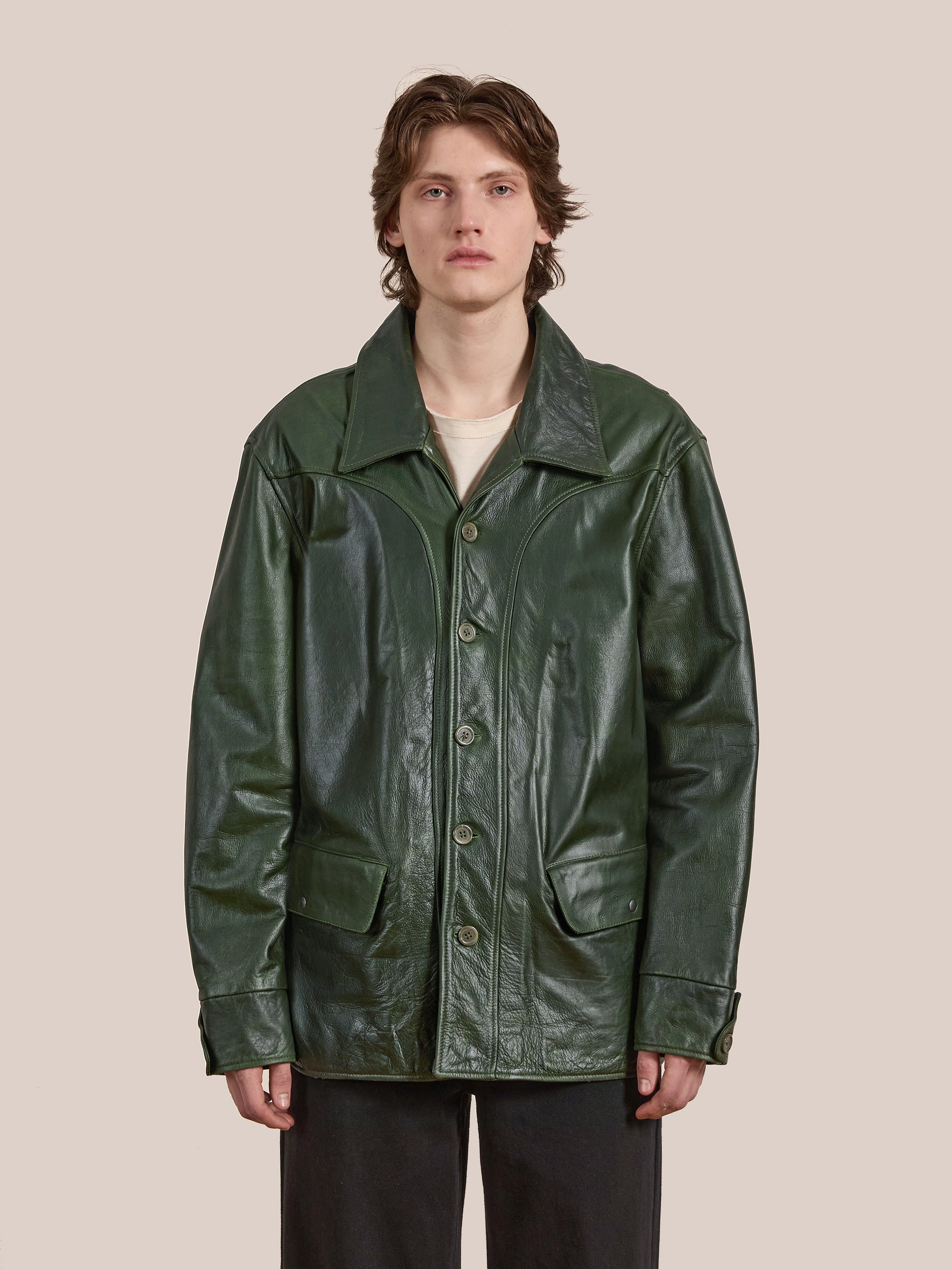 A person in the FOUND Leather Field Jacket in emerald green, made from ethically sourced leather, layered over a white shirt and standing against a plain light background.