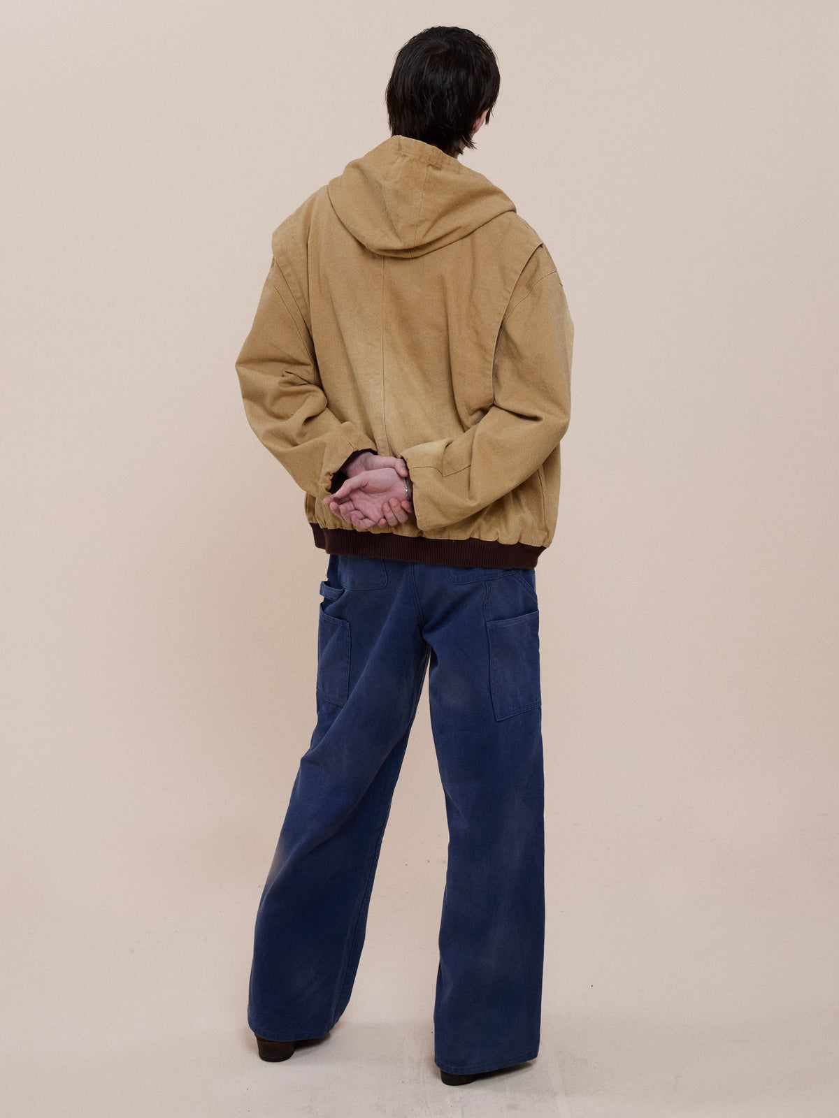 A person stands facing away, hands clasped behind their back, wearing the FOUND Faded Canvas Work Hoodie and loose blue pants against a plain beige background.