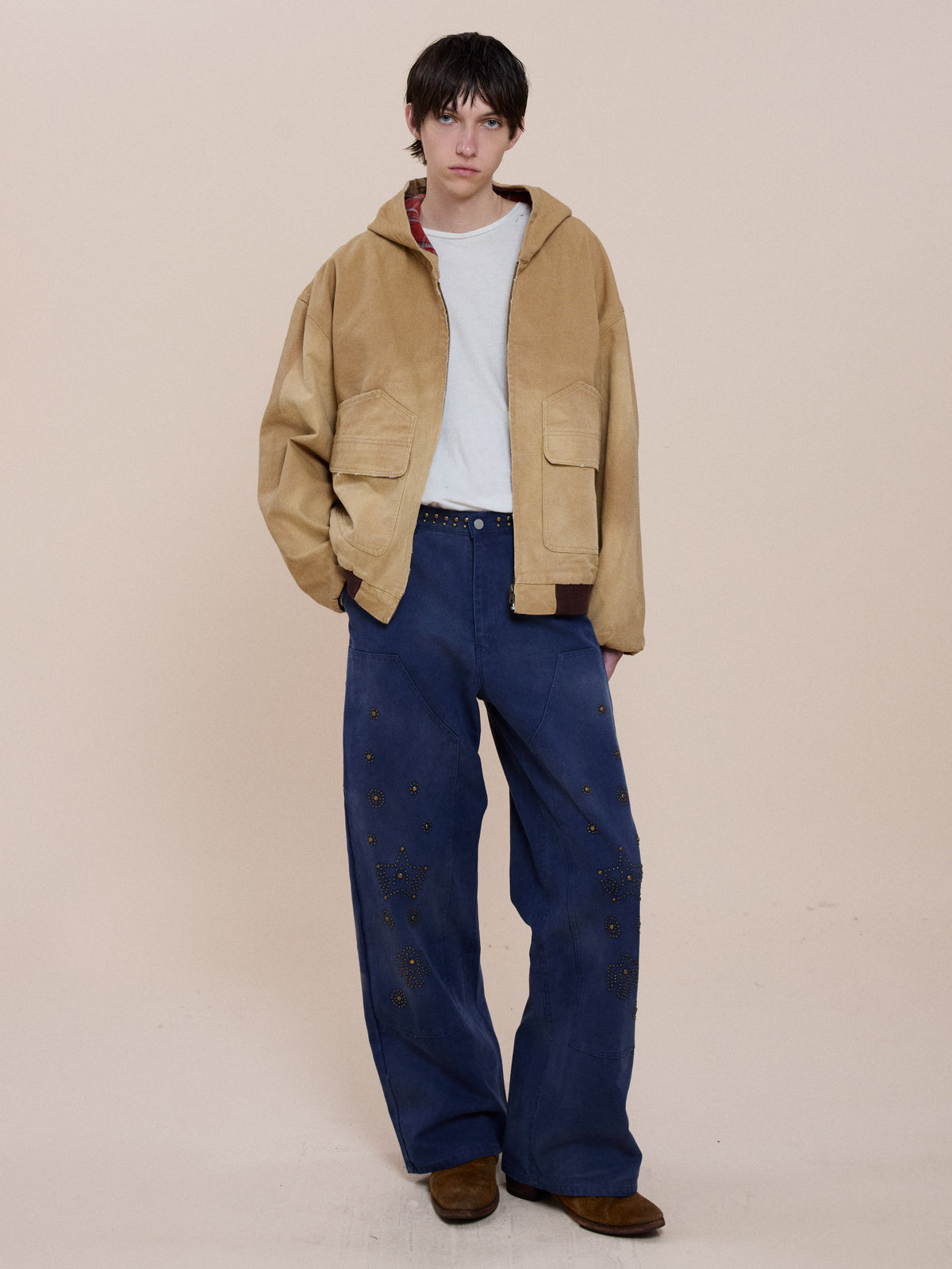 Person stands against a plain background wearing the FOUND Faded Canvas Work Hoodie, white t-shirt, embellished loose blue pants, and brown shoes.
