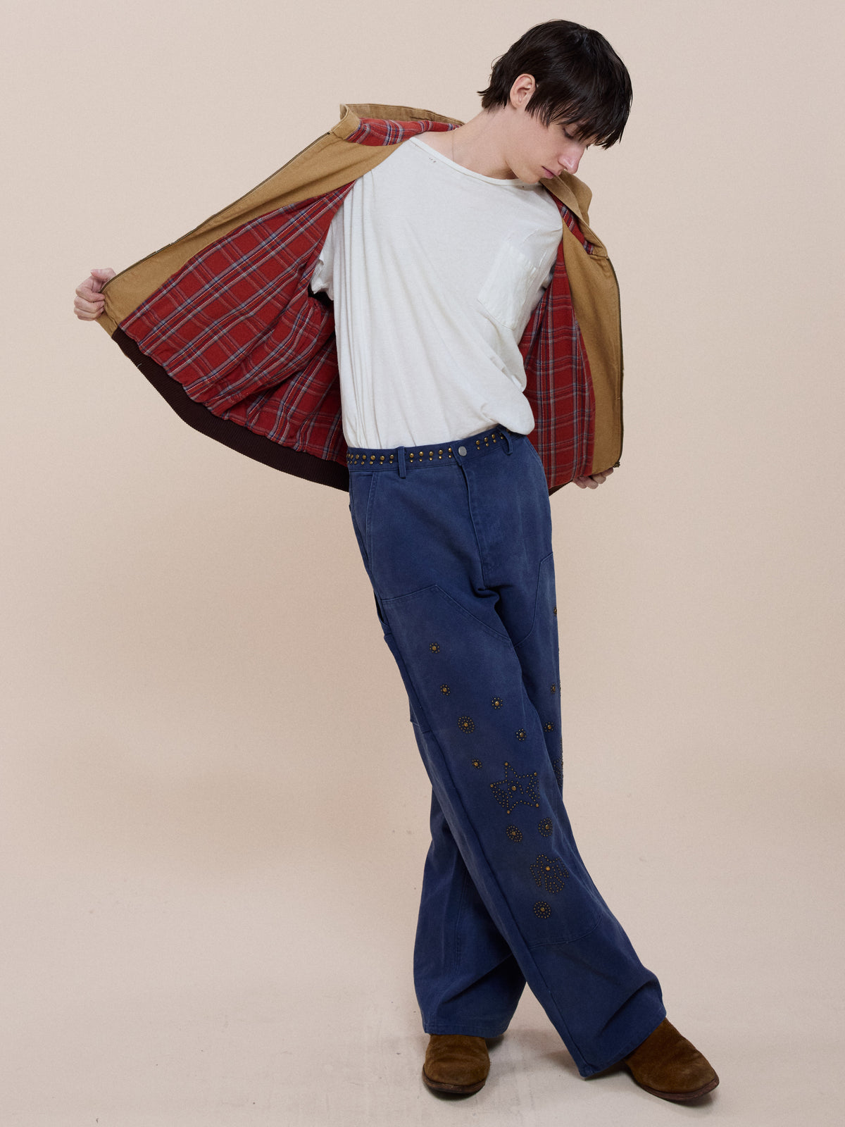 Model reveals the plaid lining of the Faded Canvas Work Hoodie by FOUND, styled with a white tee, embellished blue wide-leg pants, studded belt, and brown shoes on a beige background.
