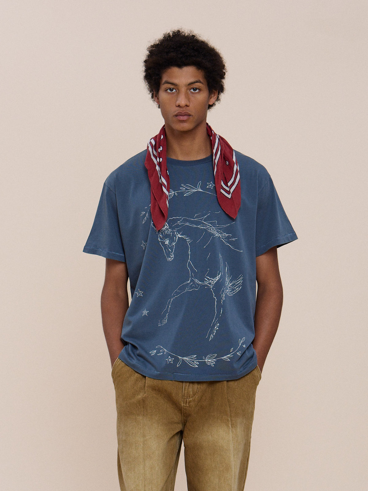 A person wears the FOUND Faded Horse Trot Tee, tan pants, and a red scarf, standing against a plain beige background.