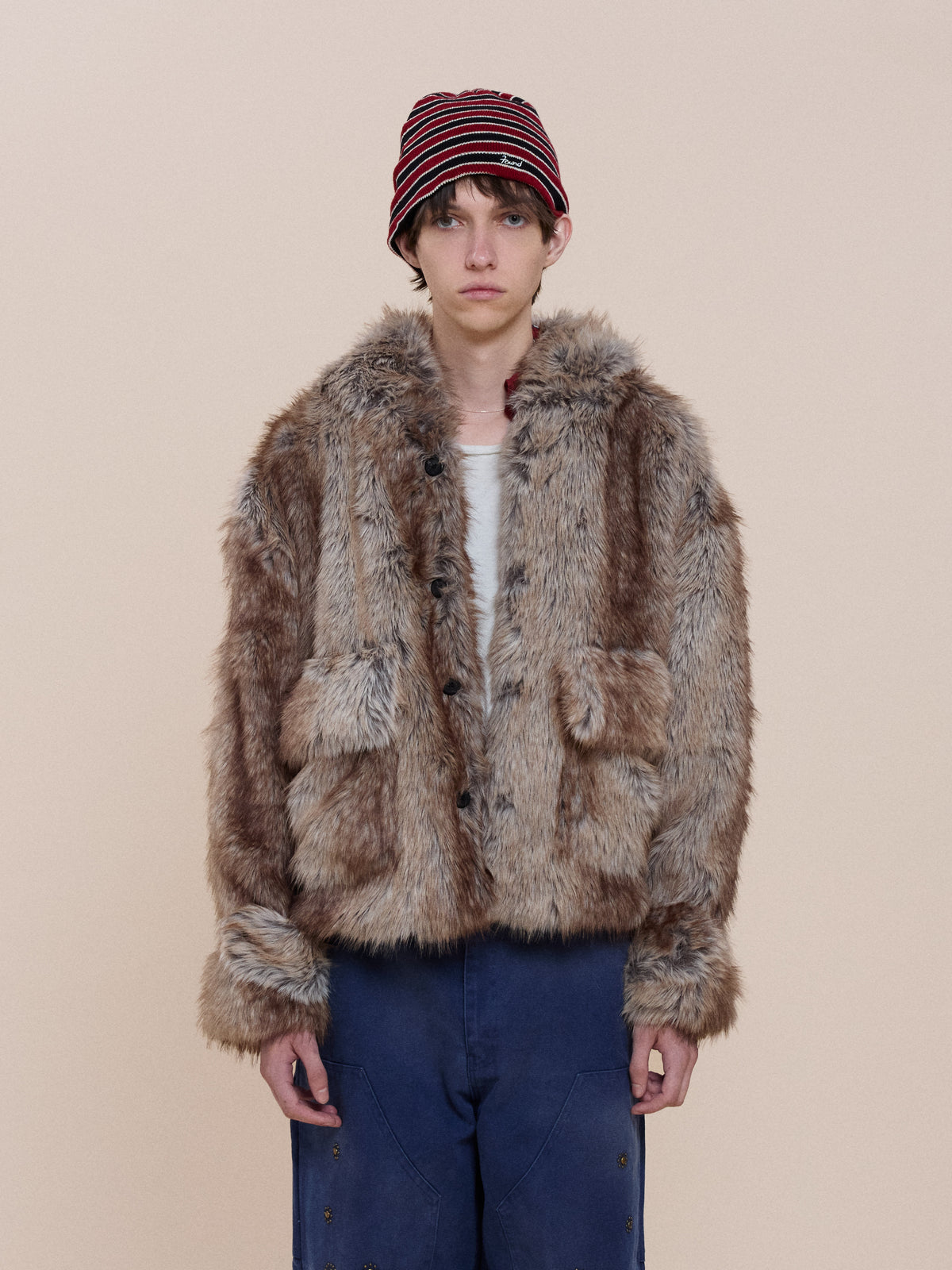 A person in a striped red and black beanie, FOUND Faux Fur Hoodie, white shirt, and blue pants stands against a plain beige background.