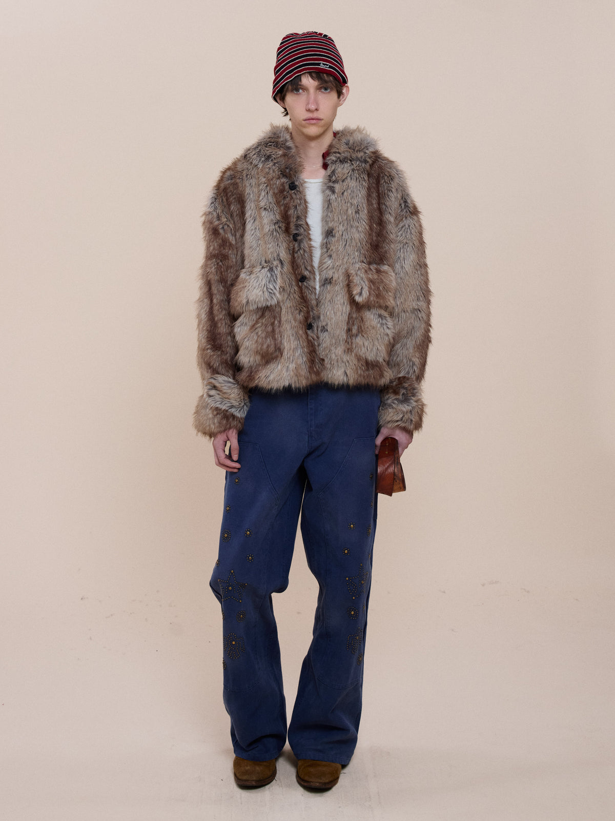 Person stands against a plain background wearing FOUND Faux Fur Hoodie, blue wide-leg pants with grommets, brown shoes, striped beanie, and holding a small pouch.