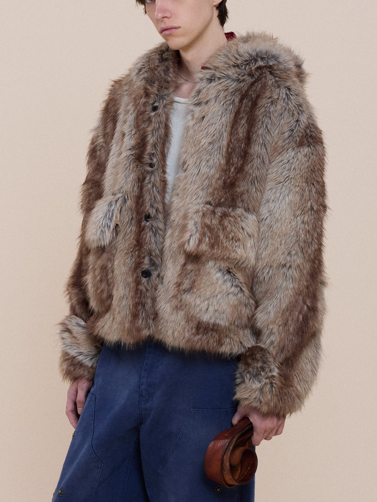 Model wears the Faux Fur Hoodie by FOUND over a white shirt and blue pants, holding a small brown accessory against a beige background.