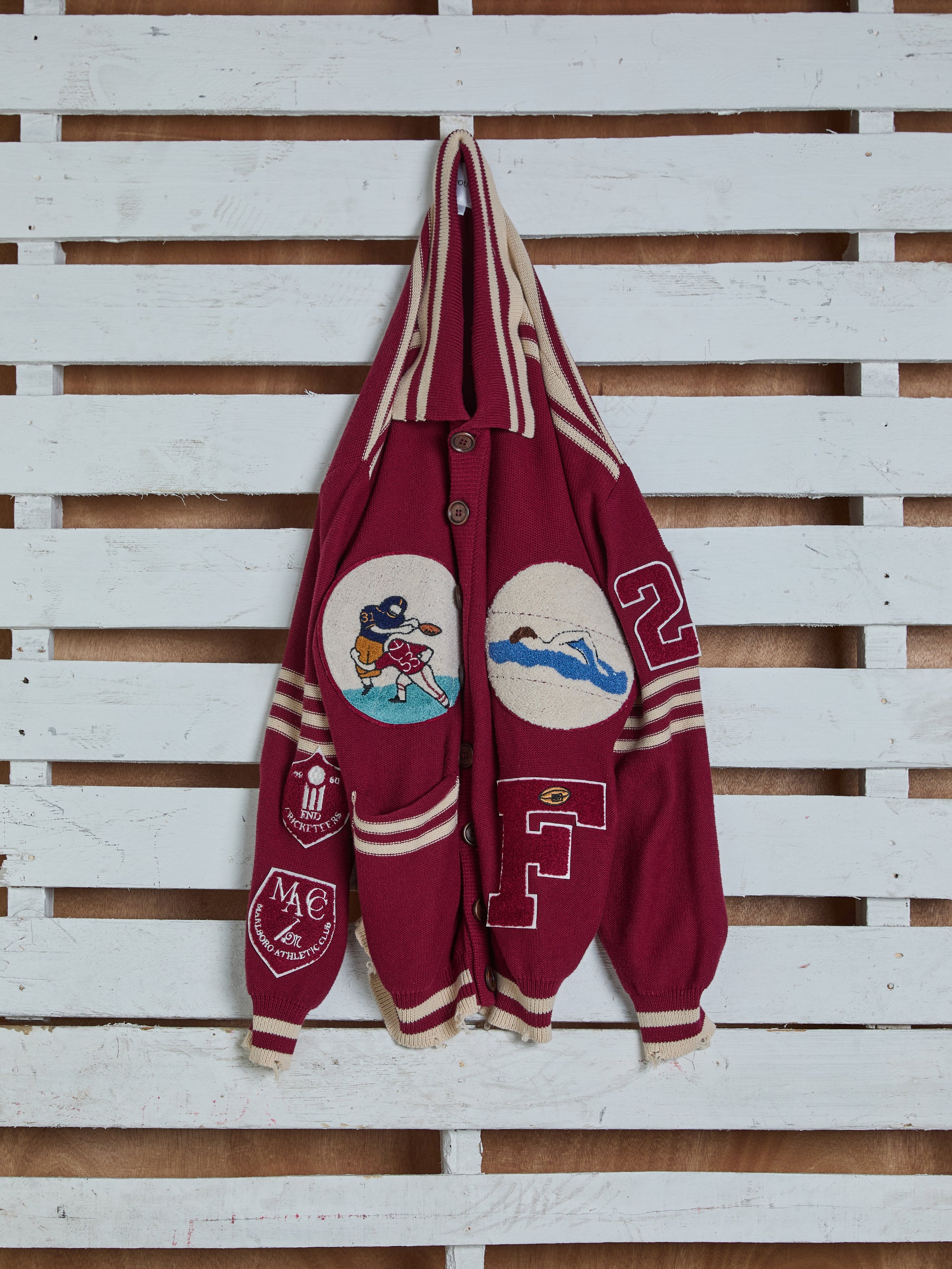 The Fin Varsity Patch Collared Cardigan by Found, a red jacket with tan stripes, hangs on a white wooden wall. It features cartoon patches, the letter "F," and numbers "22" on the right sleeve, perfectly capturing that classic Ivy League vintage vibe.