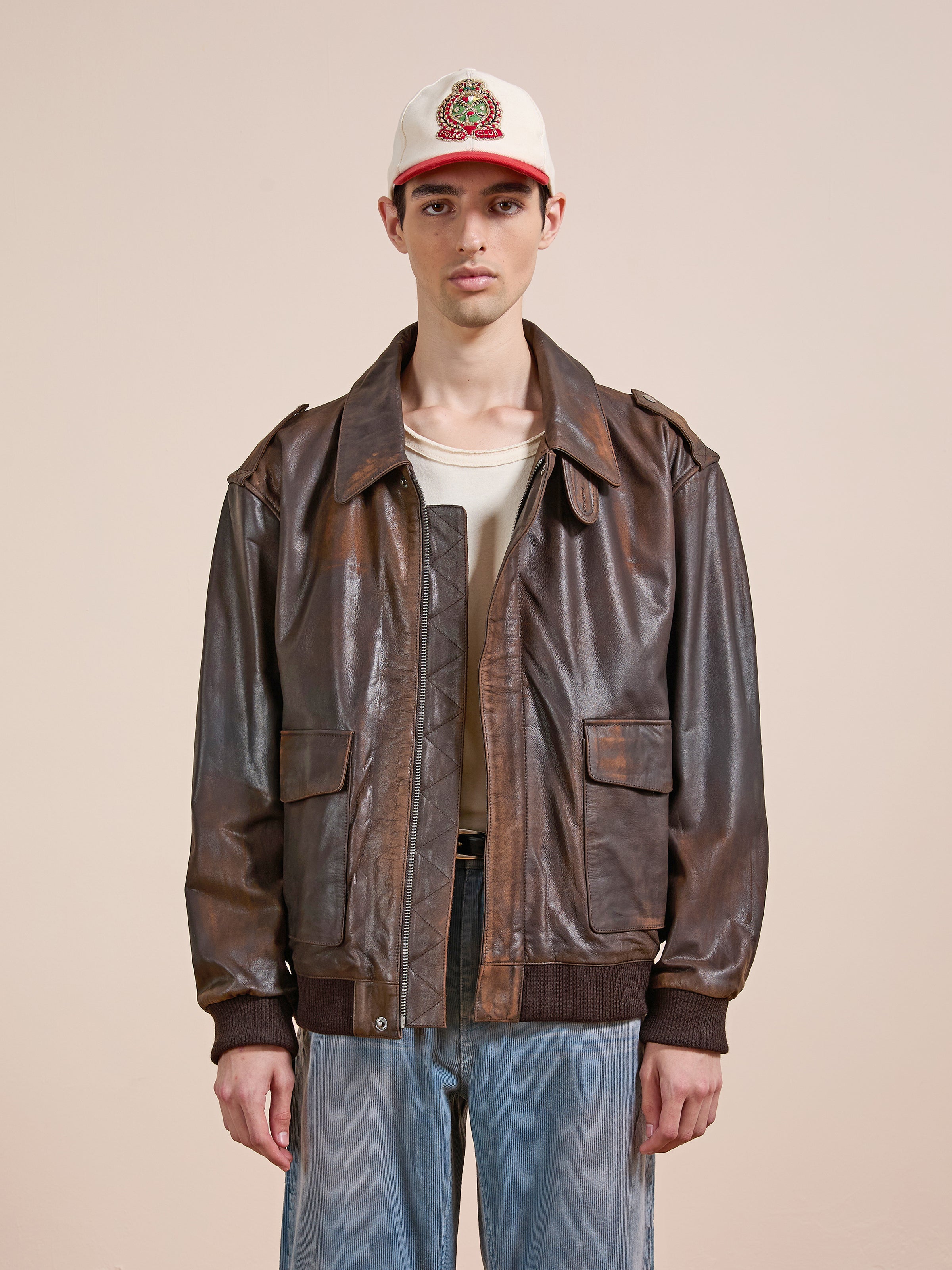 A person wears the FOUND Distressed Leather Flight Jacket over a beige shirt, paired with a white cap and light blue jeans, standing against a plain beige background.