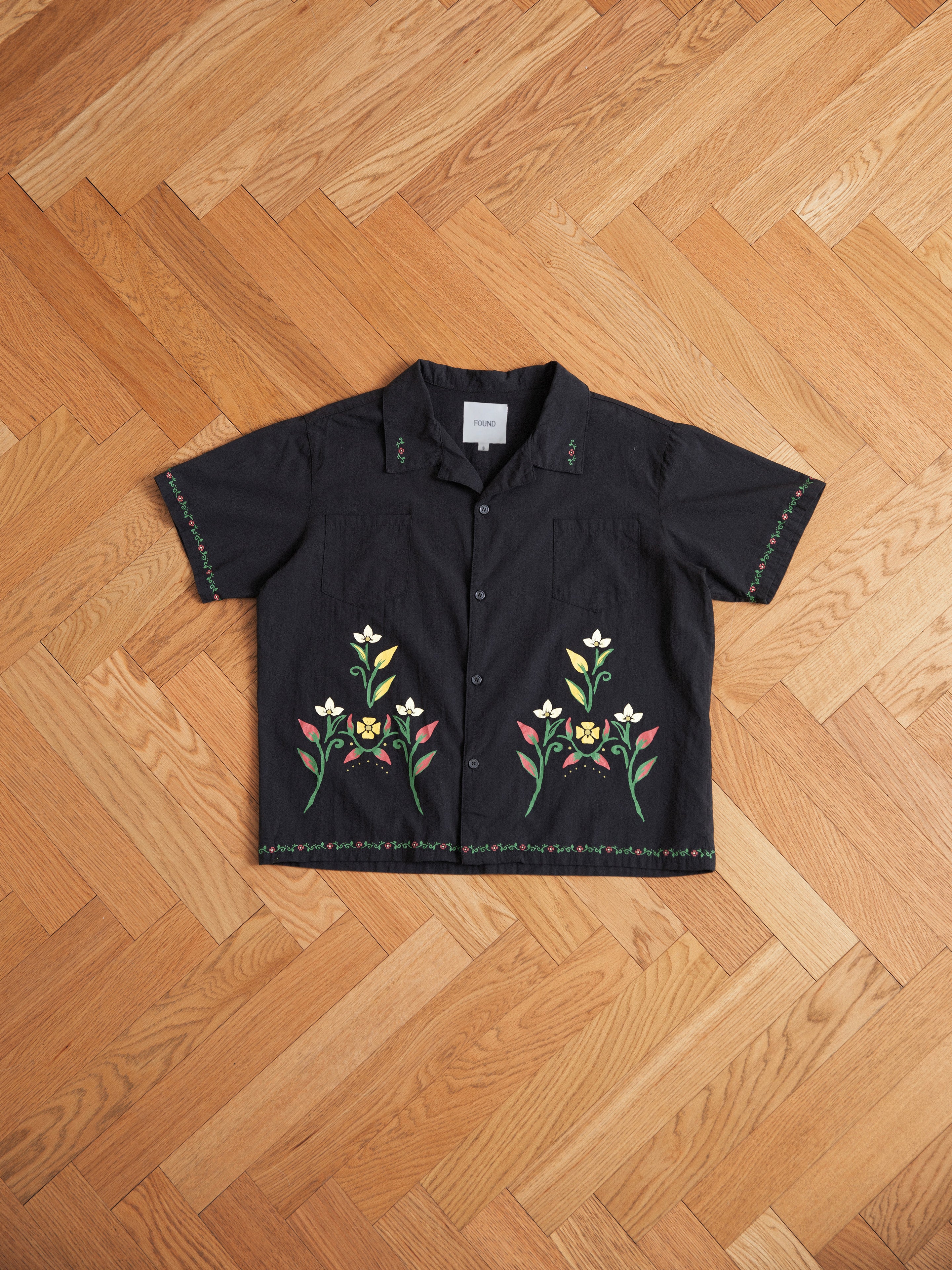 A black Floral Camp Shirt from Profound with short sleeves and floral embroidery on the front and sleeves, crafted from 100% cotton. Its relaxed fit makes it perfect for any casual occasion, displayed beautifully on a wooden herringbone floor.