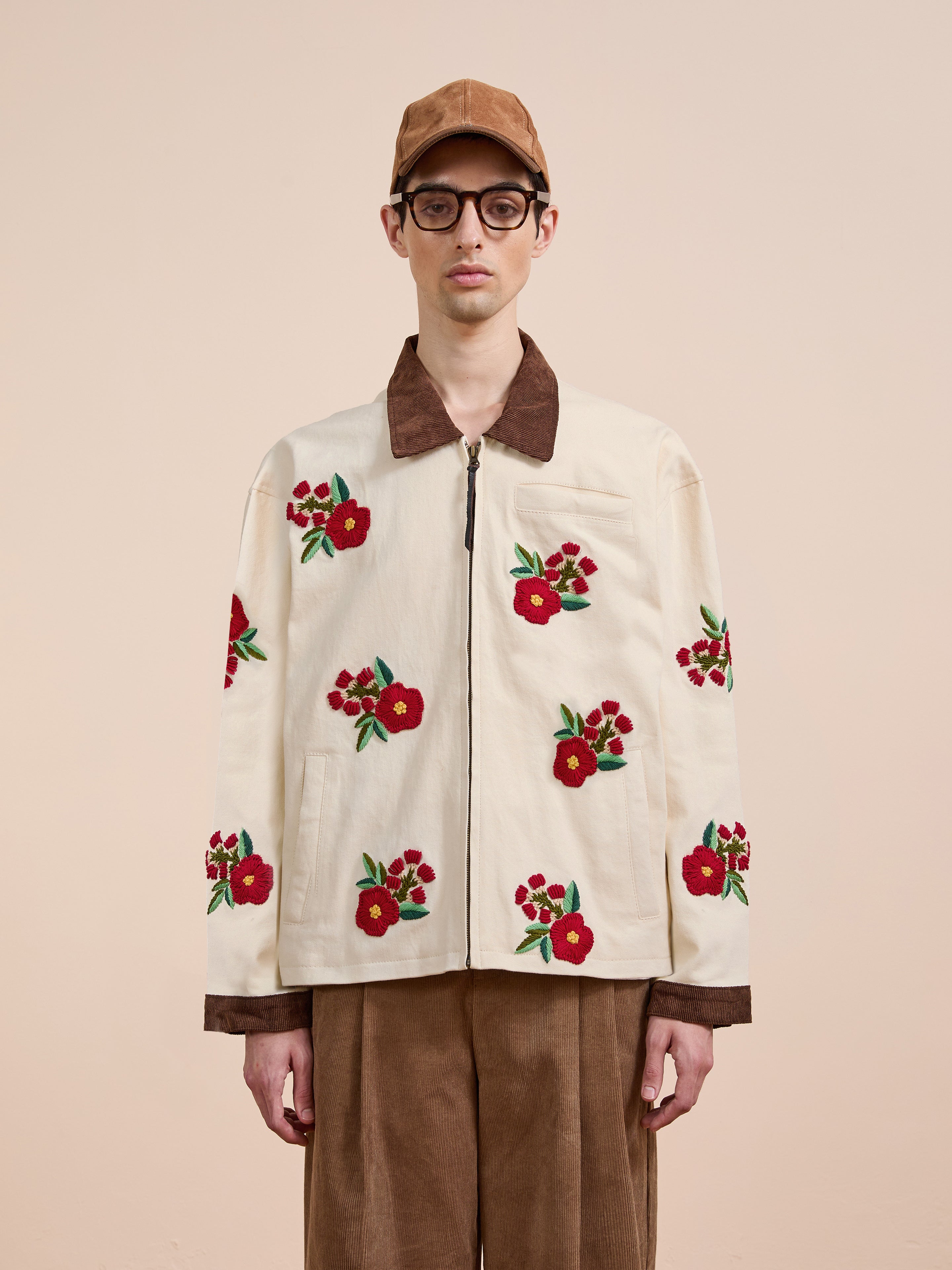 A person wearing a tan cap, glasses, brown corduroy pants, and the Found Floral Hand Embroidered Work Jacket handcrafted in Pakistan stands against a plain beige background.