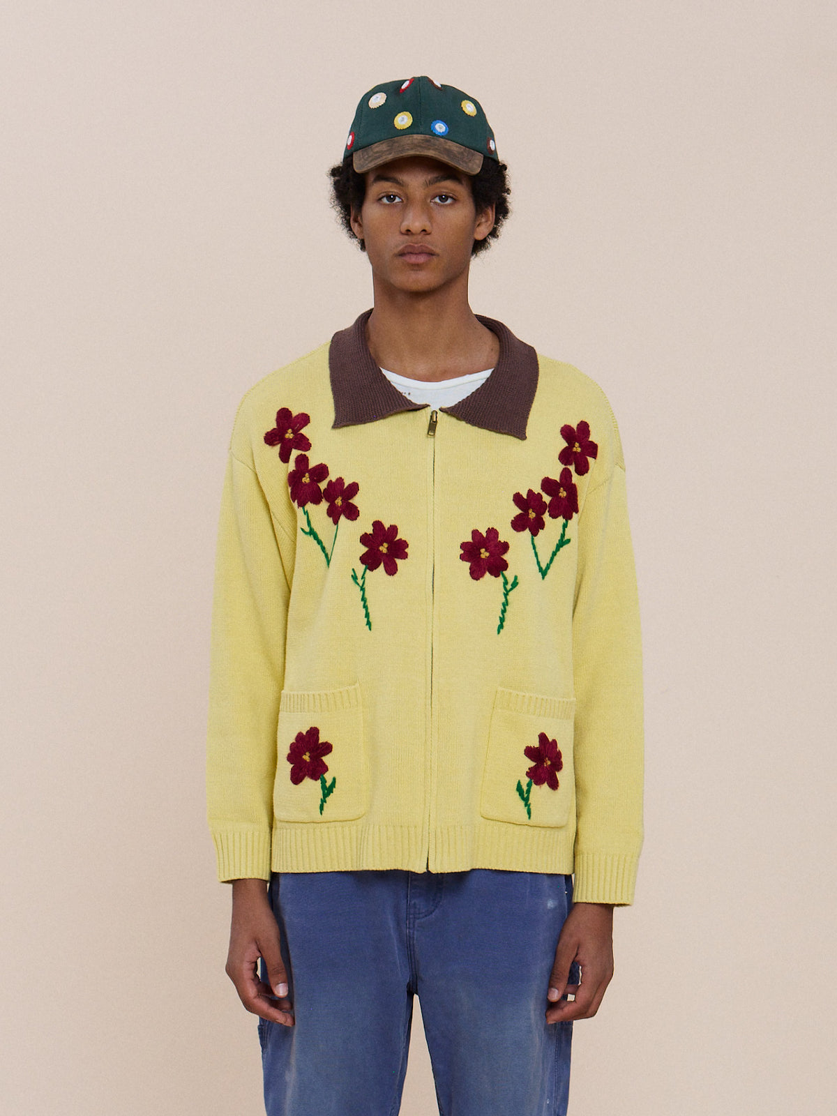 Model wears a green cap, blue jeans, and the FOUND Floral Sun Knit Sweater—yellow with red flower designs—against a plain beige background.