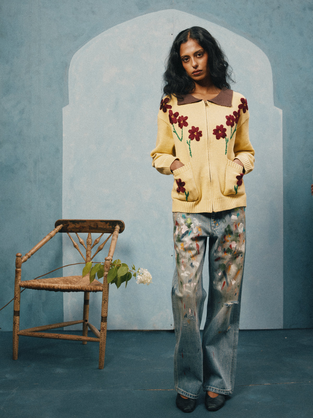Indoors, a person in the FOUND Floral Sun Knit Sweater and paint-splattered jeans stands by a rustic chair with a flower, against a blue textured backdrop.