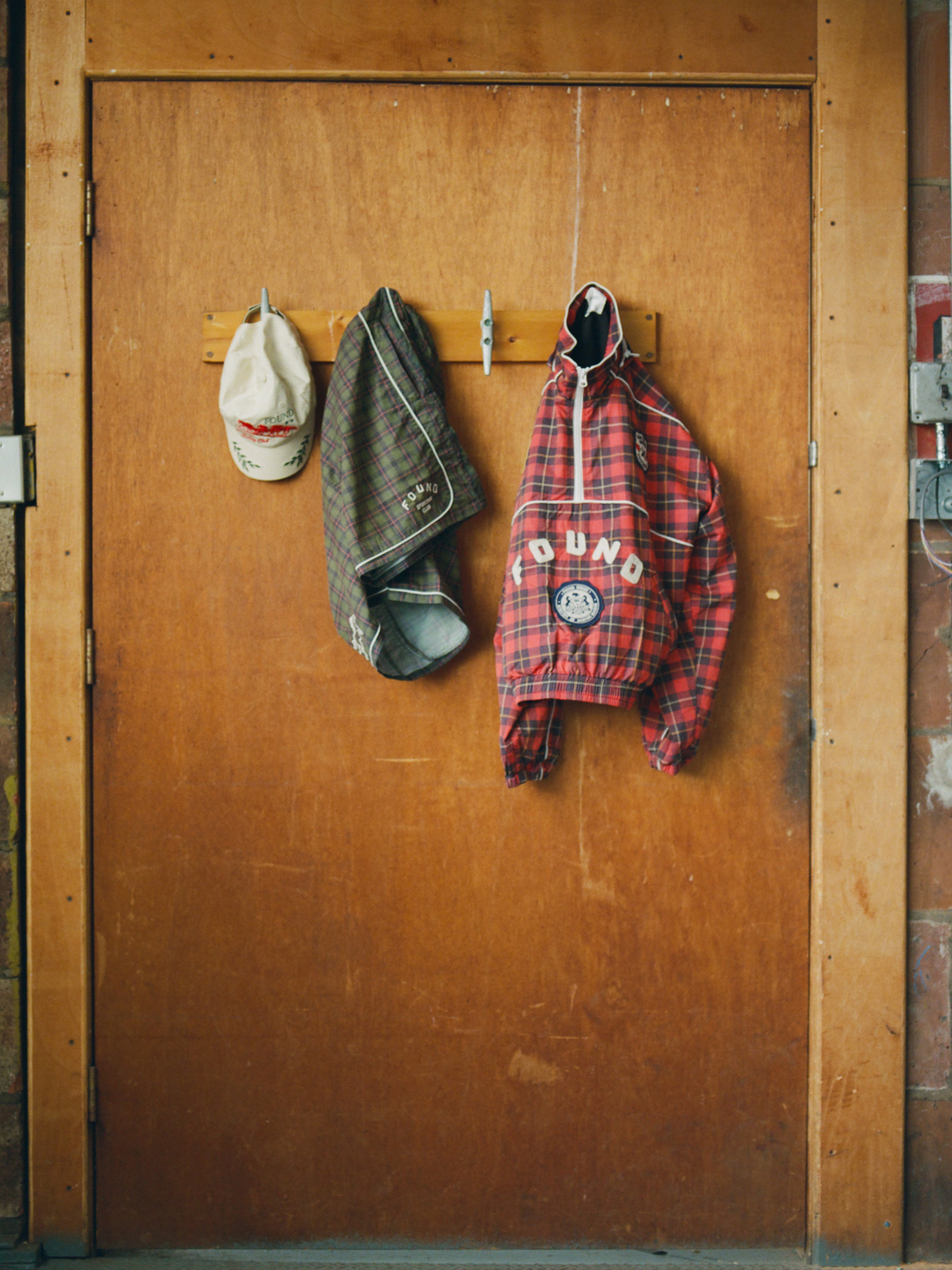 Beige cap, brown plaid shirt, and Found Forest Plaid Track Shorts with a unisex relaxed fit hang on hooks attached to a wooden door.