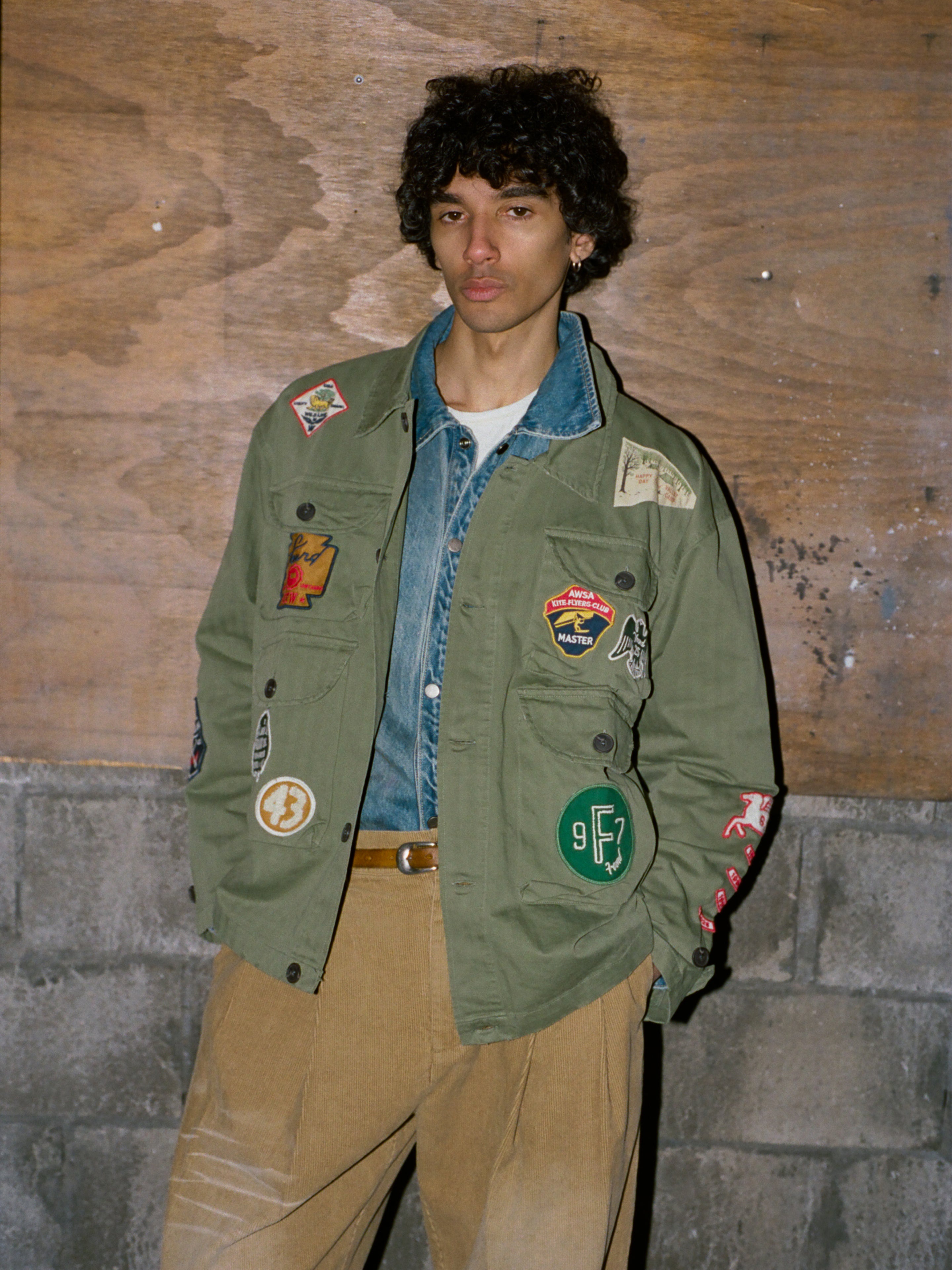A person with curly hair stands against a wooden wall, wearing the FOUND Herringbone Utility Patchwork Jacket over a denim jacket and tan pants.