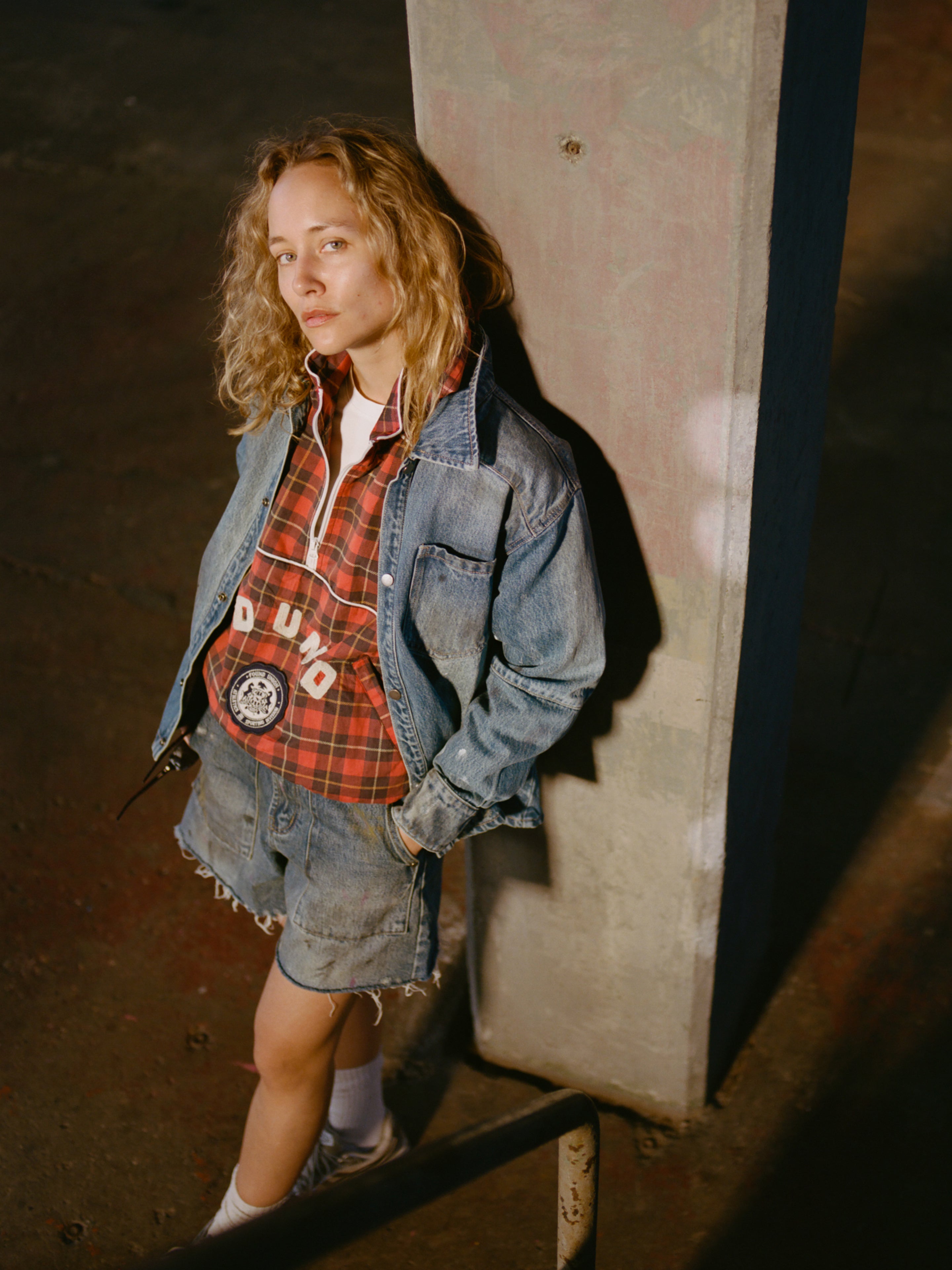 A person with shoulder-length blonde hair wears the FOUND Western Denim Painter Shirt under a denim jacket and shorts, standing by a concrete pillar in dim lighting.