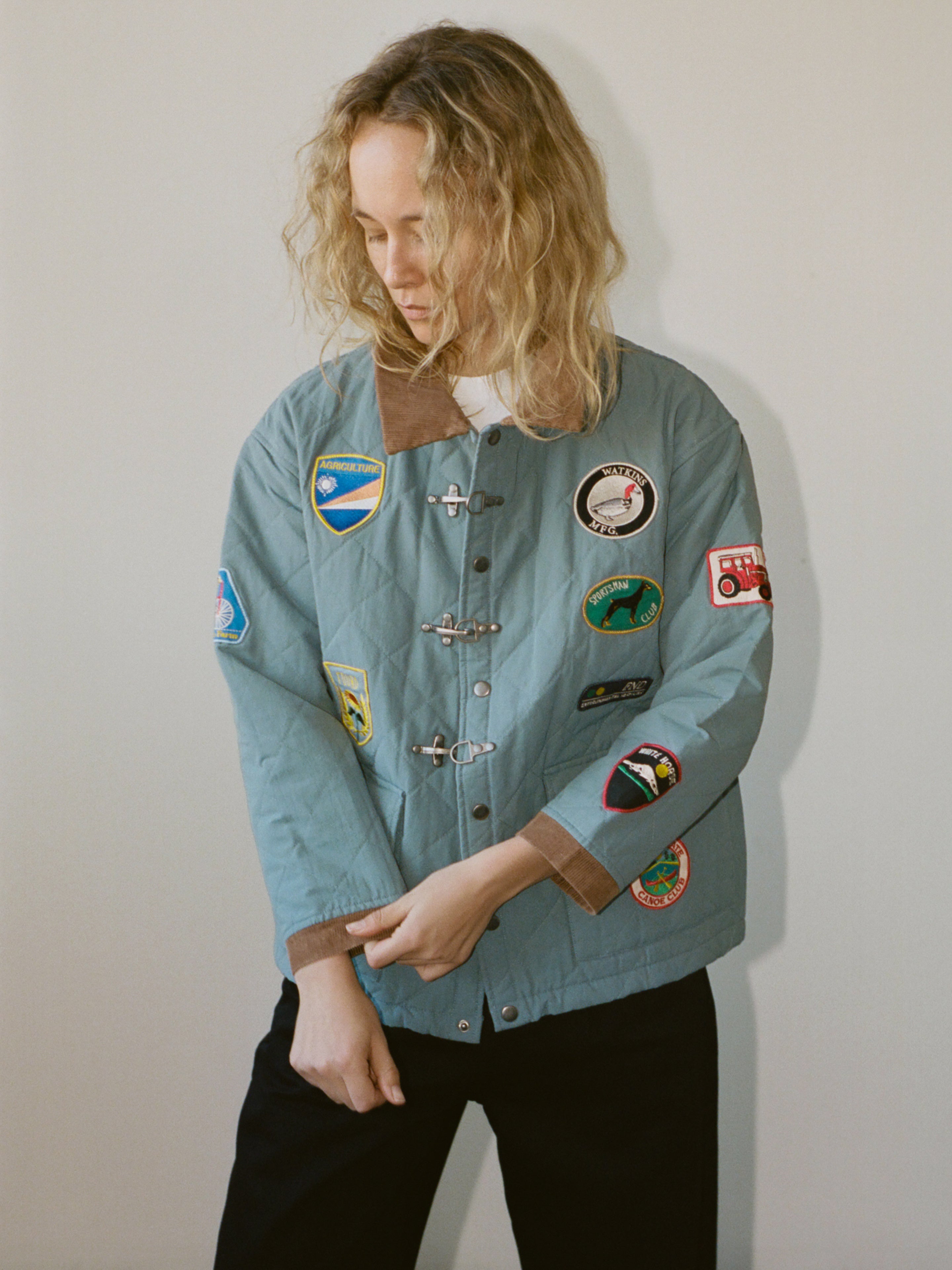 A person with wavy blonde hair wears the FOUND Farmstead Quilt Patch Jacket, a light blue piece featuring colorful patches, and stands against a plain light background.