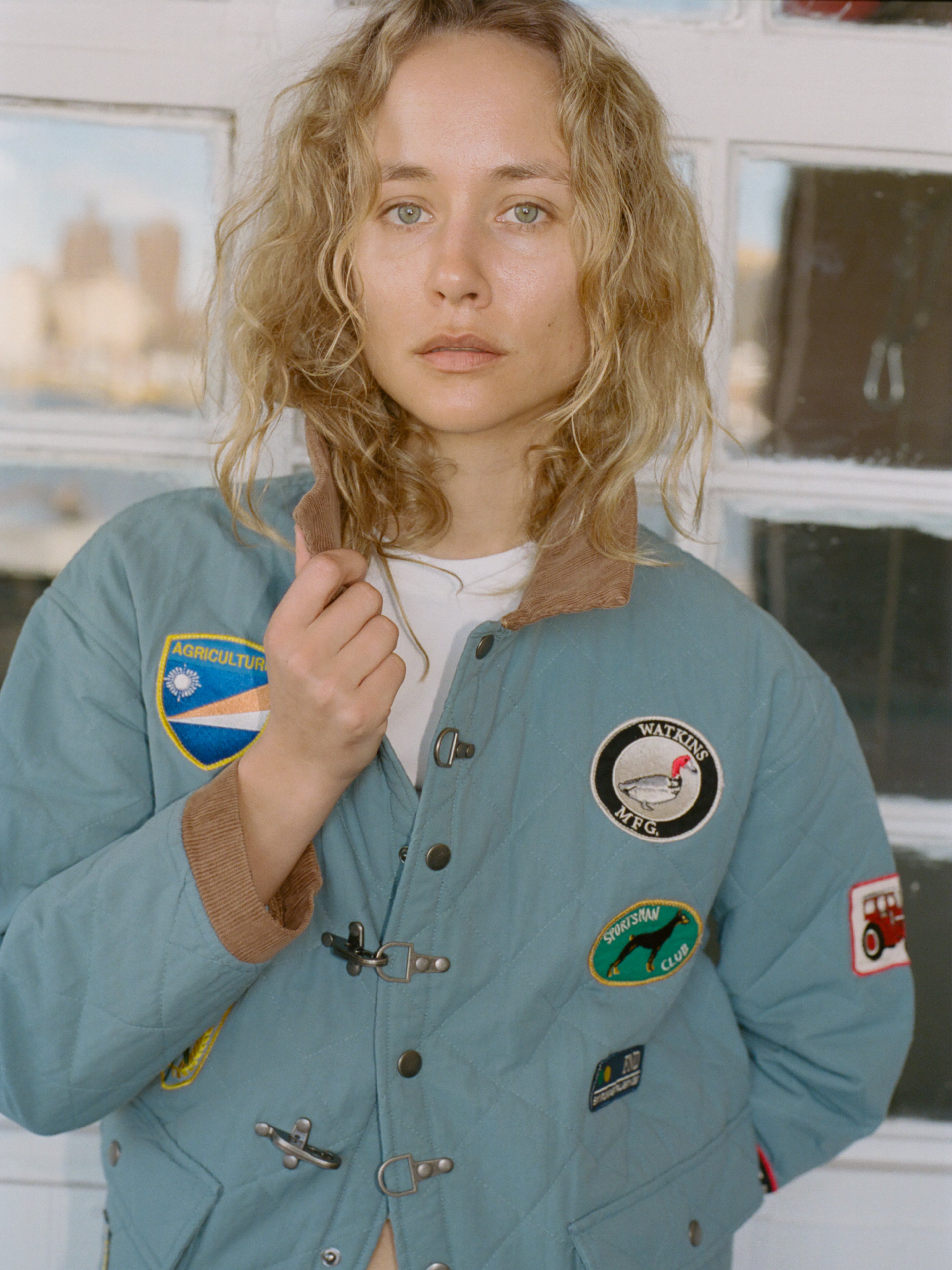 A person with curly blonde hair wears the FOUND Farmstead Quilt Patch Jacket in light blue, adorned with assorted patches, while standing before a door with windows.