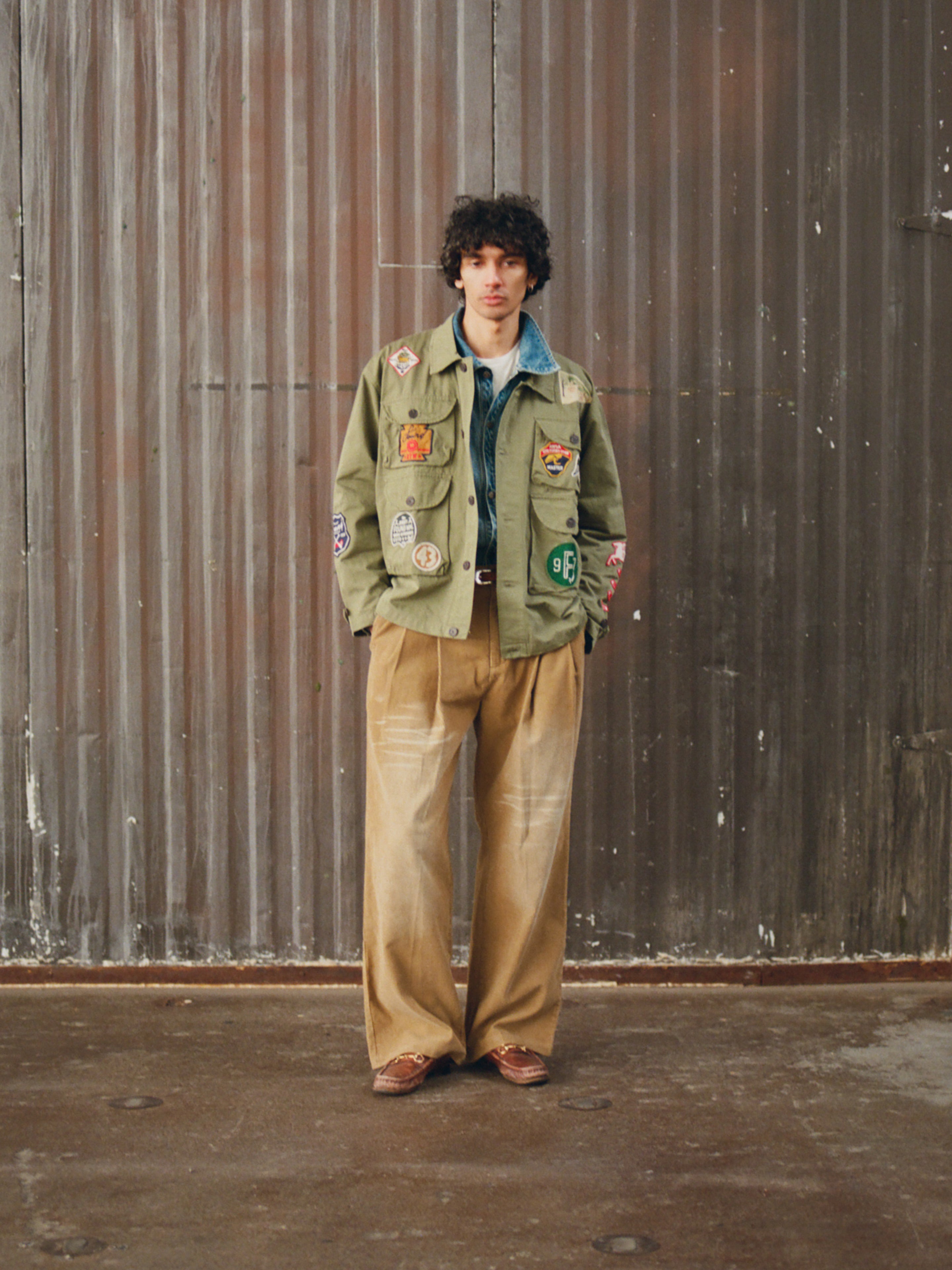 A person stands before a corrugated metal wall, wearing a green patch-covered jacket, light blue shirt, FOUND Canvas Studded Work Pants, and brown shoes.