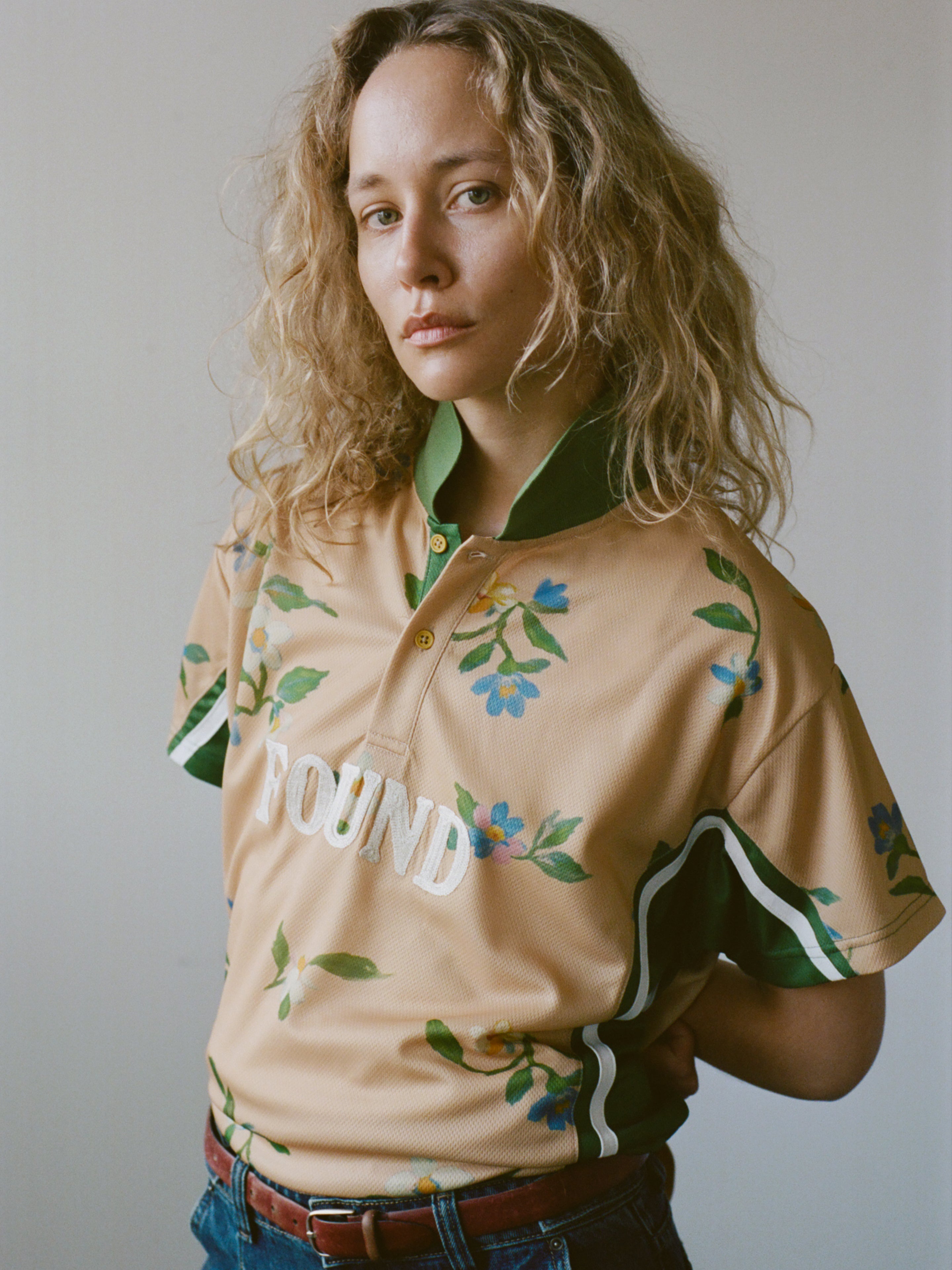 A person with curly blonde hair wears the FOUND Floral Polo Jersey featuring botanical embroidery, standing against a plain background.