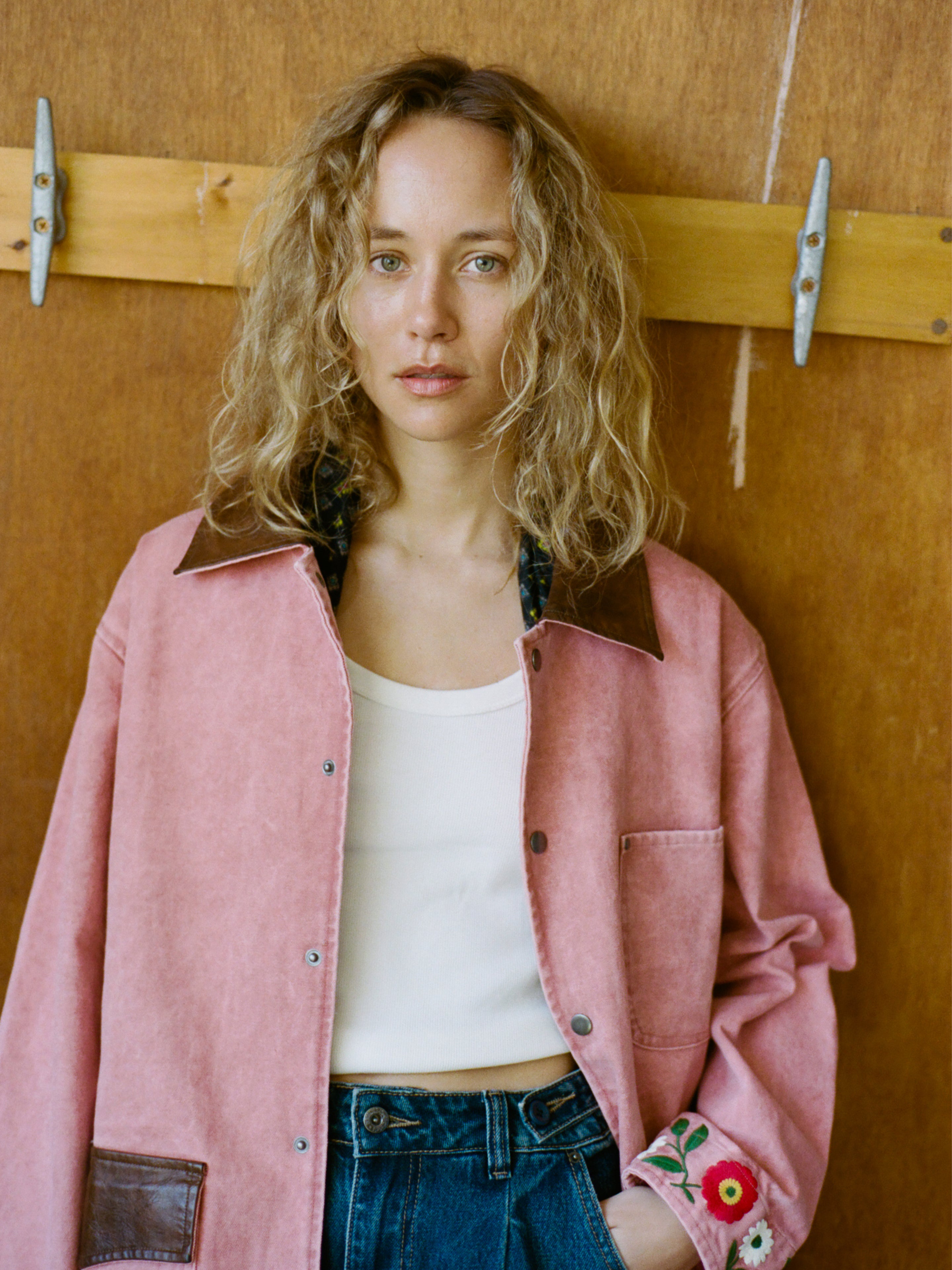 A woman with curly blonde hair models the Found Canvas Embroidered Barn Jacket over a white top and blue jeans, standing against a wooden background and looking at the camera.