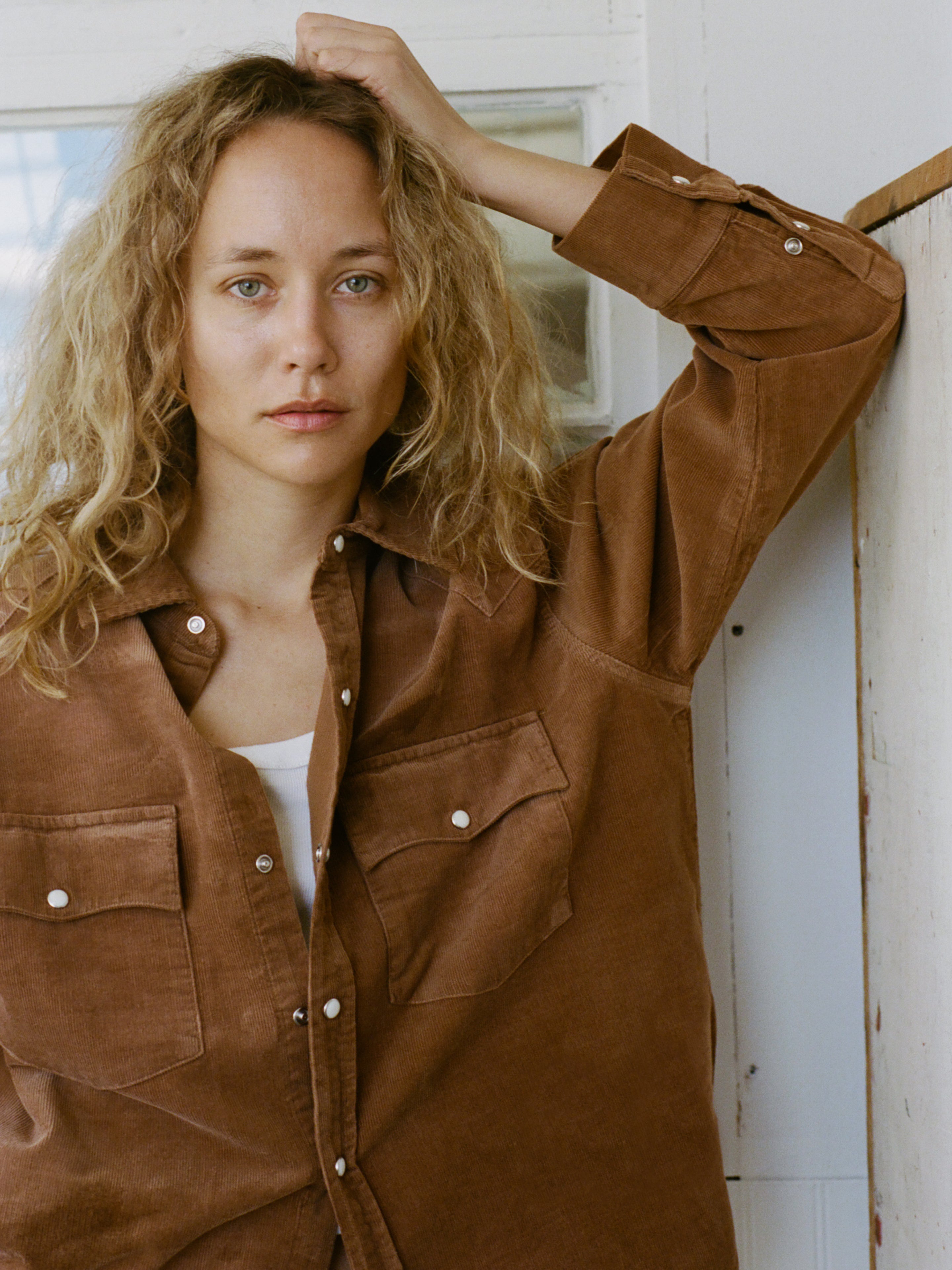 A person with long curly blonde hair wears the FOUND Western Corduroy Shirt in a unisex relaxed fit, standing indoors with one arm resting on a white wall and looking at the camera.