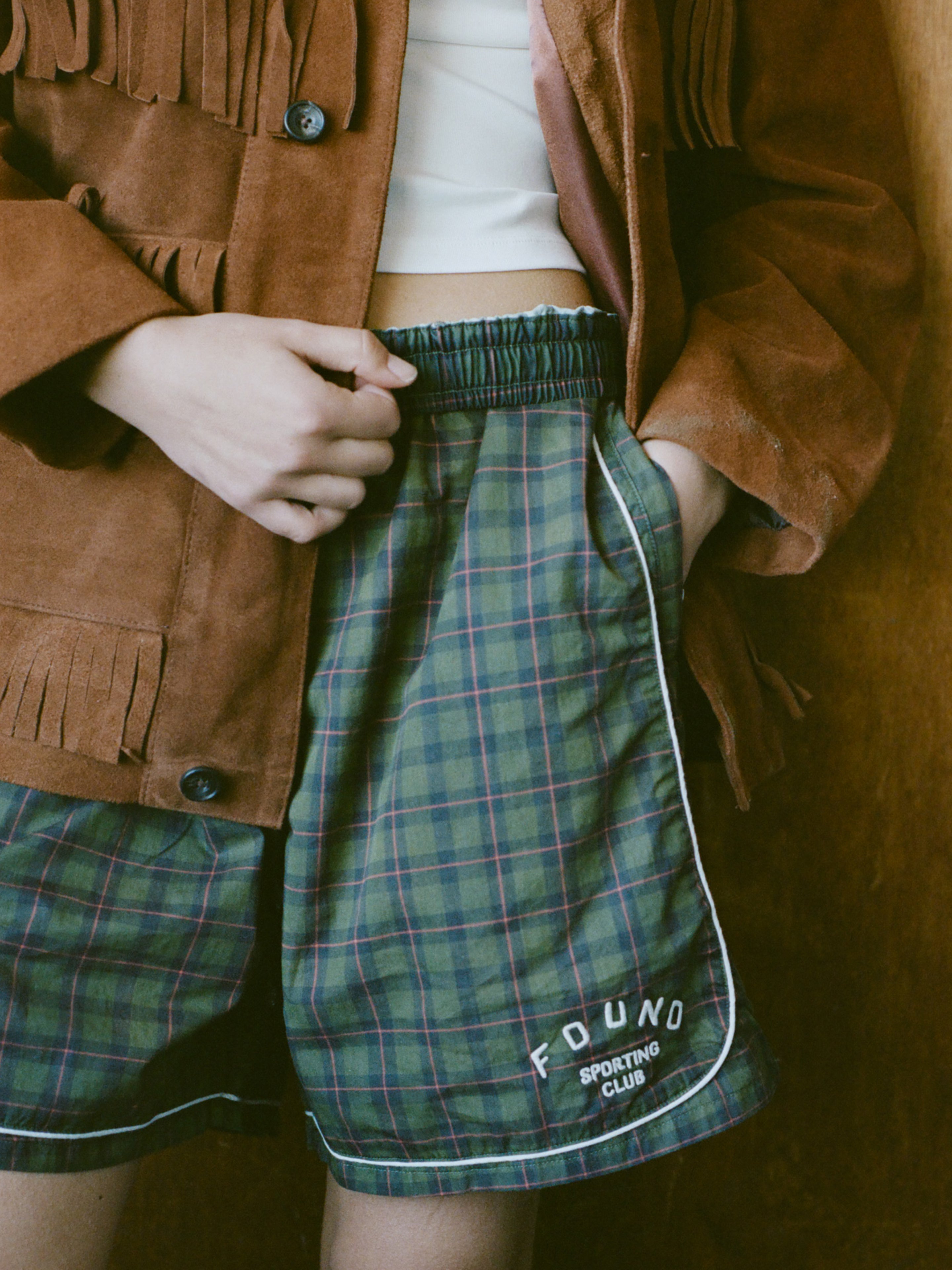 A person wearing a brown suede fringe jacket, a white crop top, and Found's Forest Plaid Track Shorts.