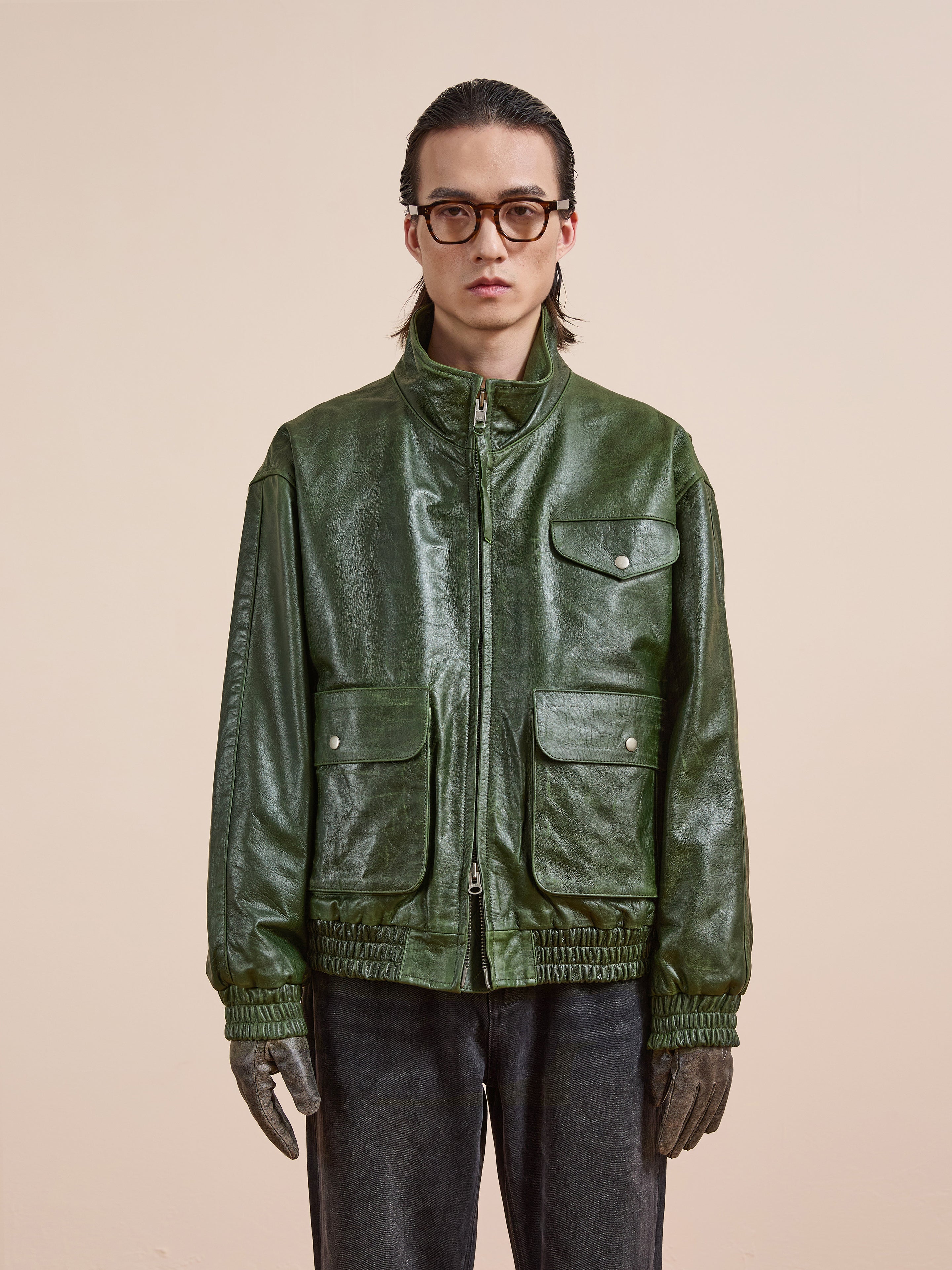 A person in a FOUND Forest Leather Bomber Jacket, dark gloves, jeans, and large glasses stands before a beige background. This genuine leather bomber offers unisex, bold style with timeless appeal.