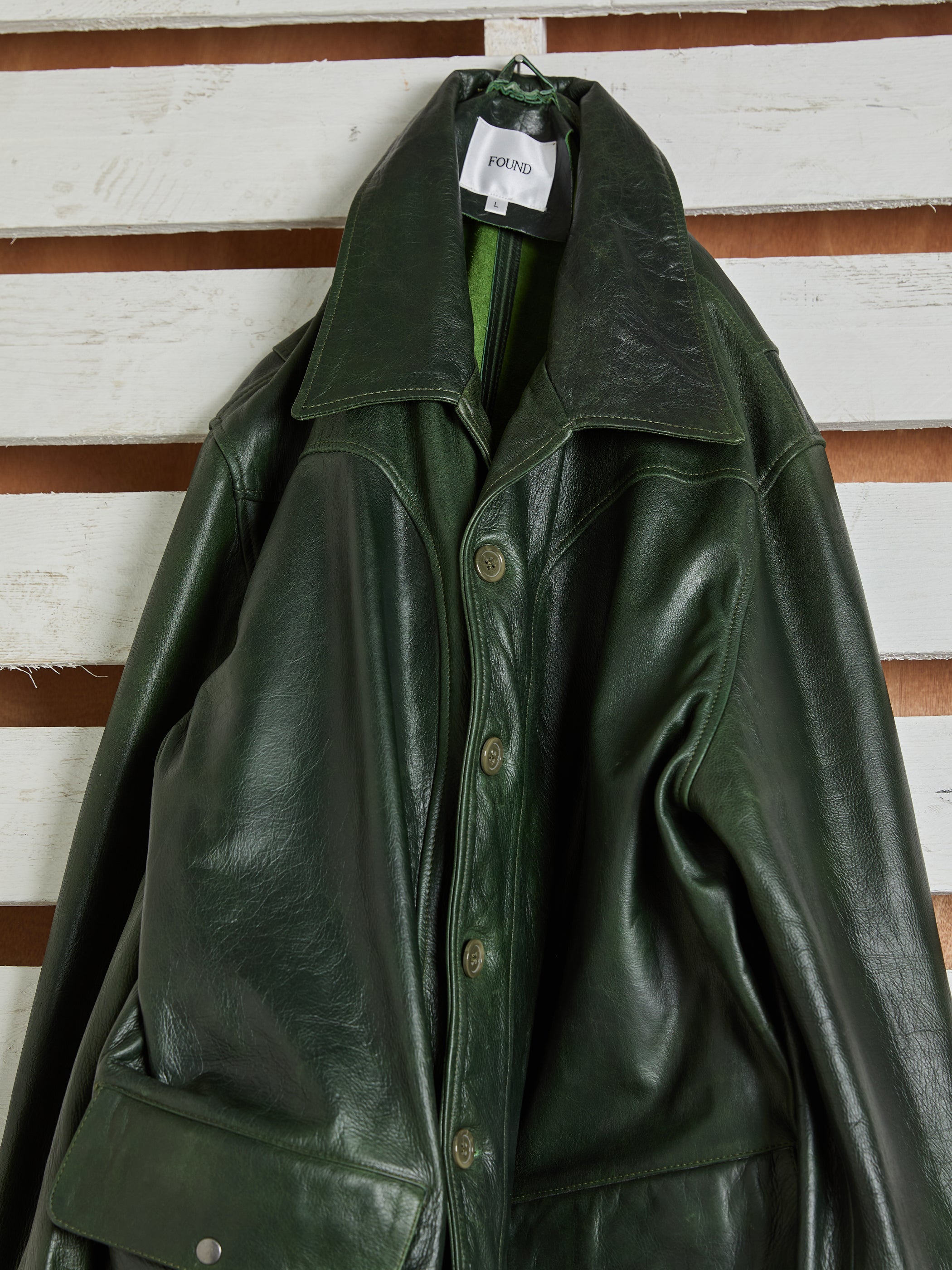 The FOUND Leather Field Jacket, crafted from hand-distressed, ethically sourced leather in emerald green, hangs on a hanger against a white wooden slat wall.