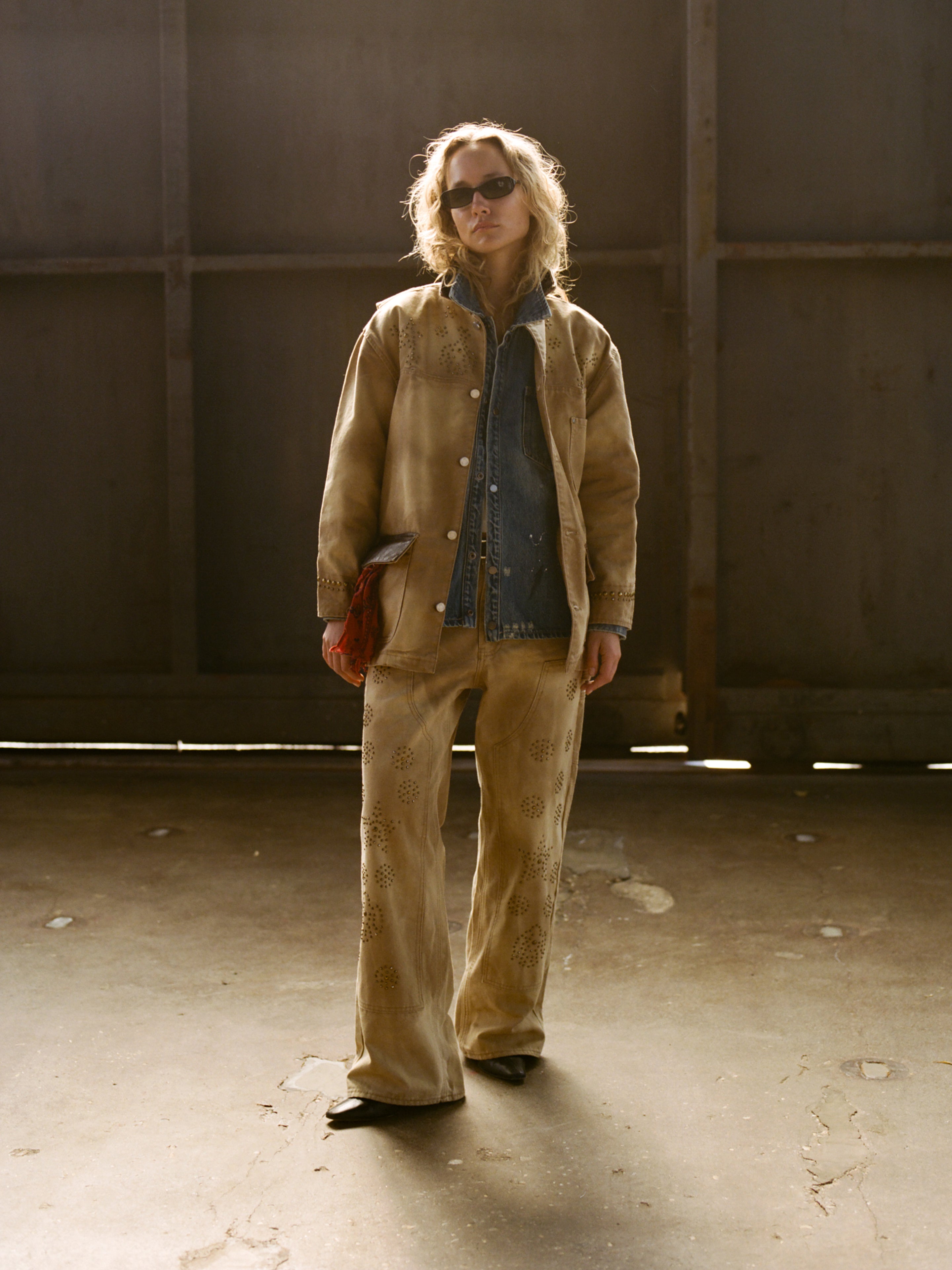 Person wearing sunglasses, a tan embroidered jacket with FOUND Canvas Studded Work Pants over a blue denim shirt, holding a red clutch, standing in a sunlit industrial setting.