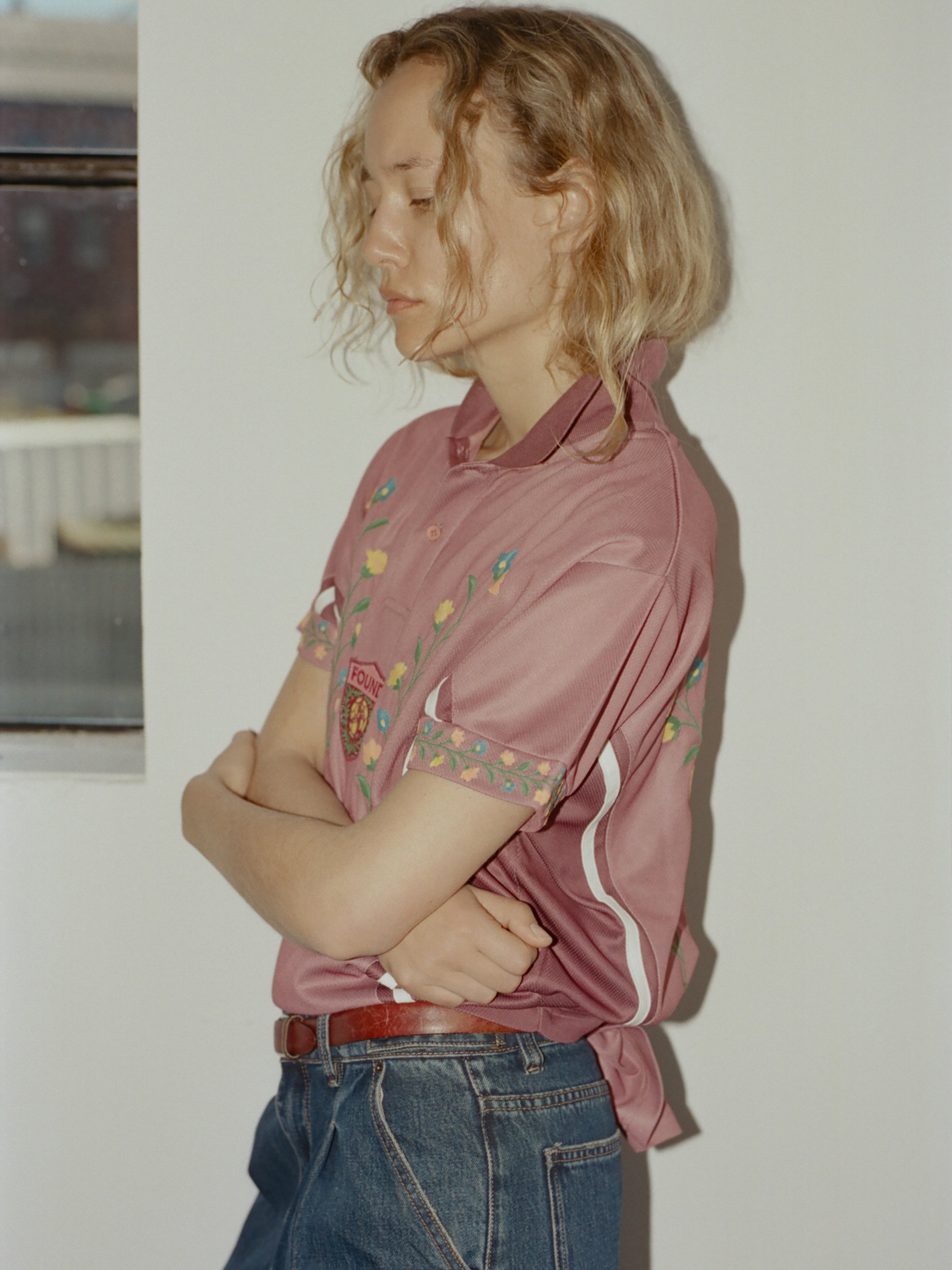 A person with shoulder-length blonde hair, wearing the FOUND Floral Polo Jersey and blue jeans, stands indoors facing sideways with arms crossed.