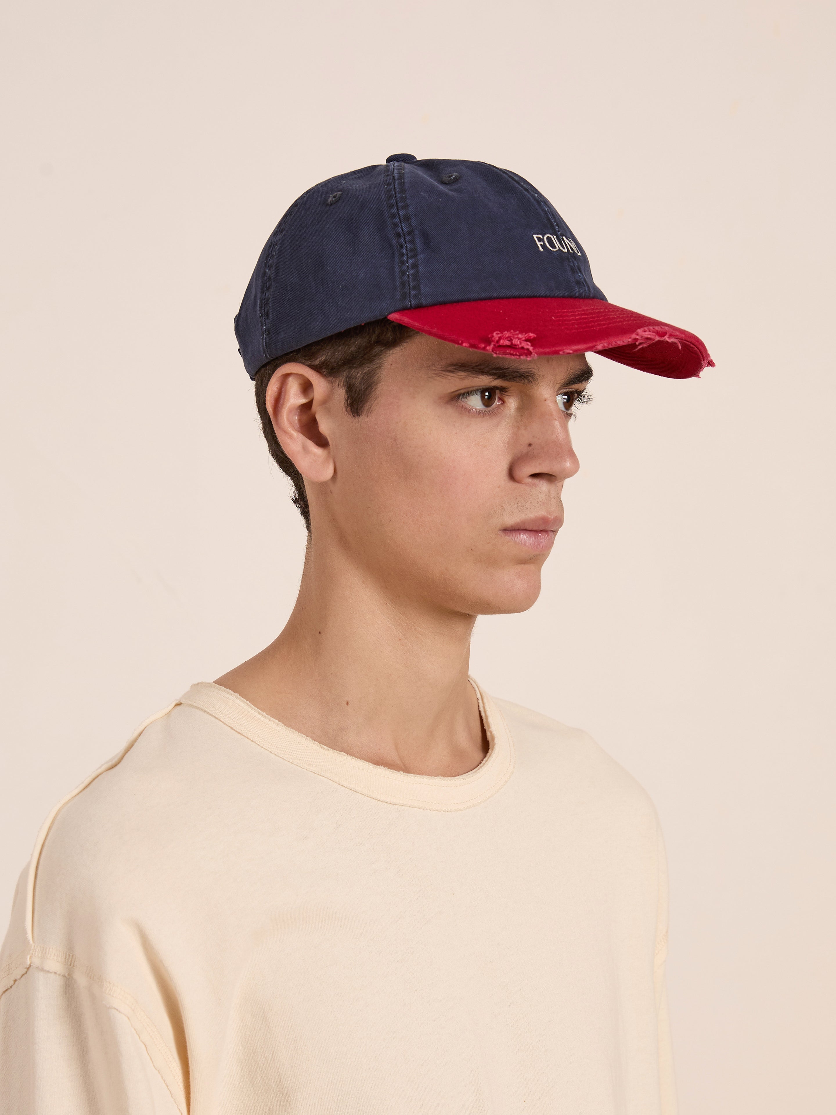 A person wearing a FOUND Two-Tone Logo Cap with a navy cotton top, adjustable Velcro back strap, distressed red brim, and a cream-colored shirt is looking to the right against a plain background.