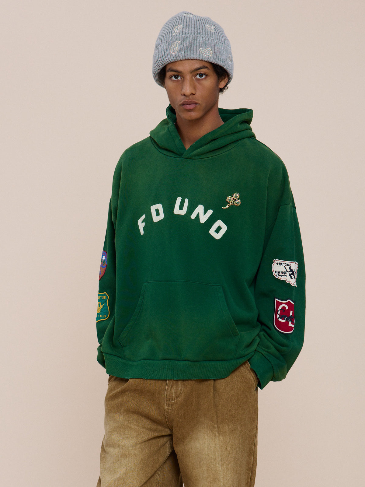 Person wears the FOUND Found Logo Patch Hoodie in vintage forest green, tan pants, and a light gray beanie, standing against a plain beige background.
