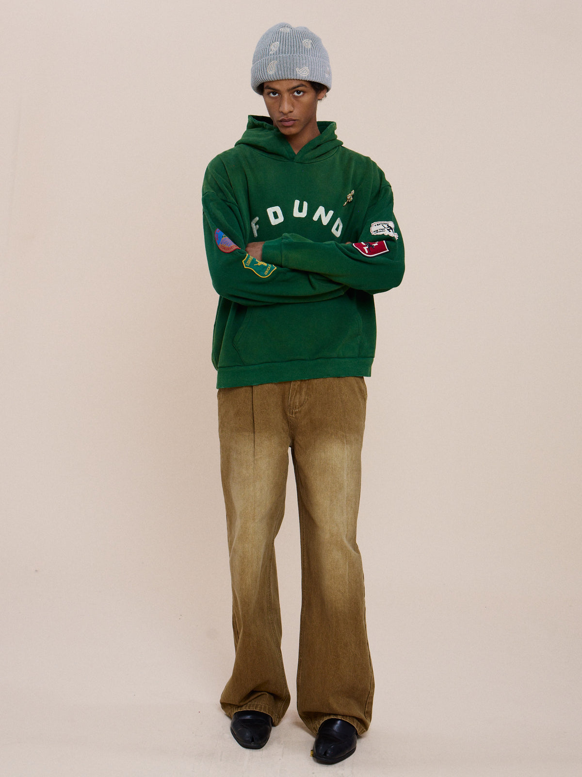 Person standing with arms crossed, wearing the FOUND Found Logo Patch Hoodie in vintage forest green, tan corduroy pants, black shoes, and a light gray knit beanie.