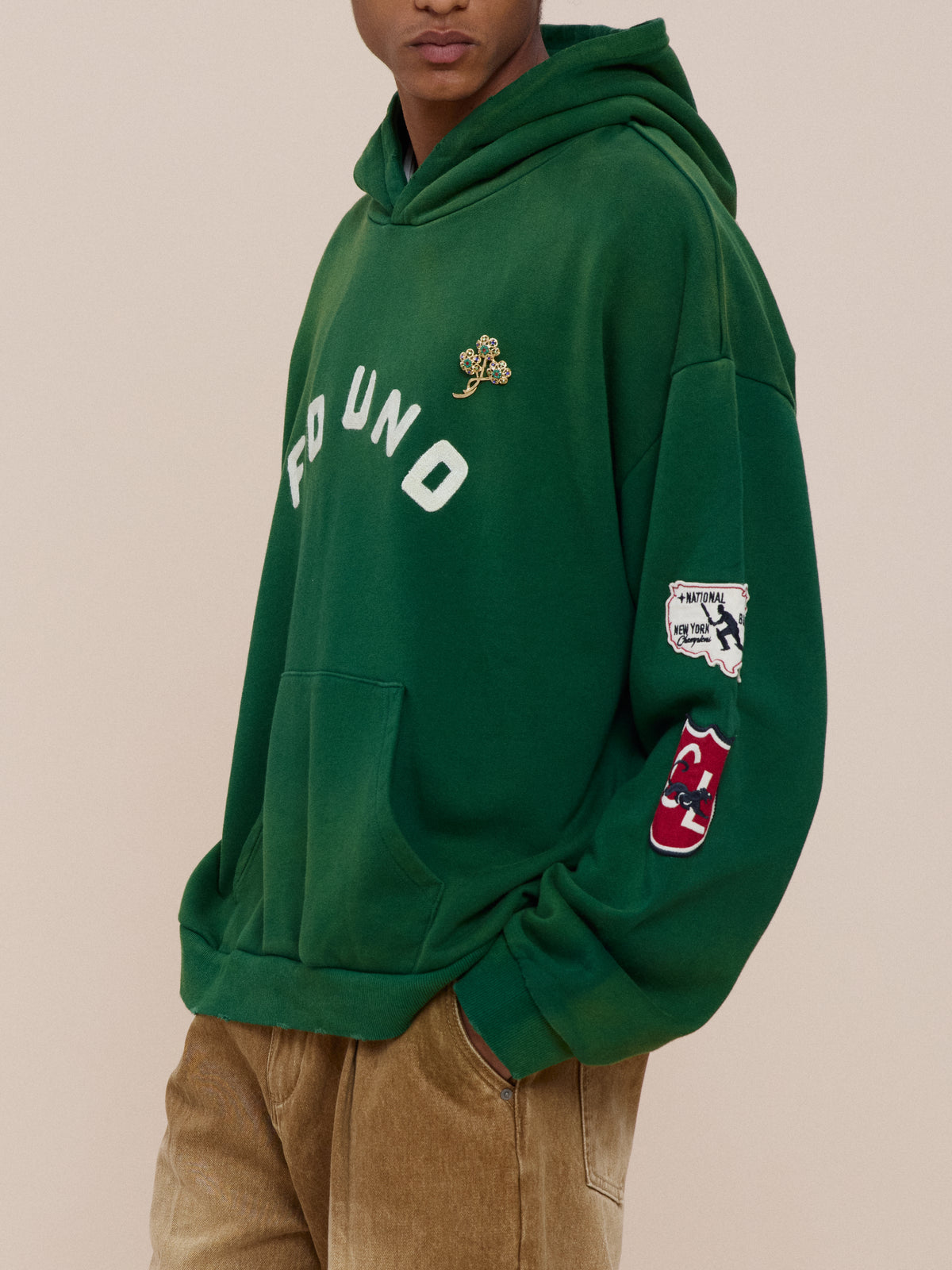 Model wears the FOUND Found Logo Patch Hoodie—vintage forest green with logo patches—and light brown pants, standing against a neutral background.