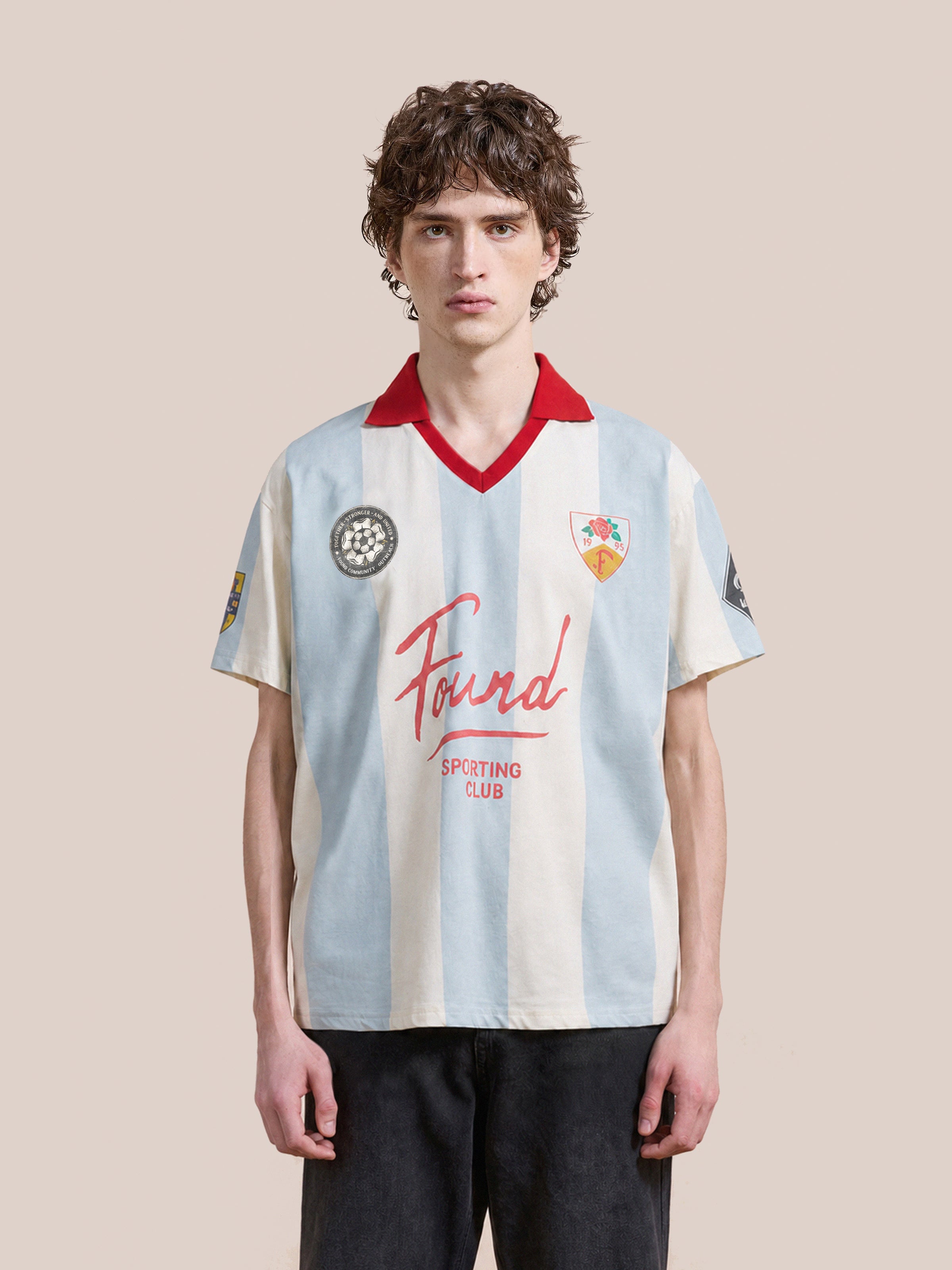 A person wearing the FOUND Found Club Short Sleeve Jersey—a unisex white and light blue striped top featuring a red collar, multiple patches, and “Found Sporting Club” text inspired by classic football shirts—stands against a plain background.