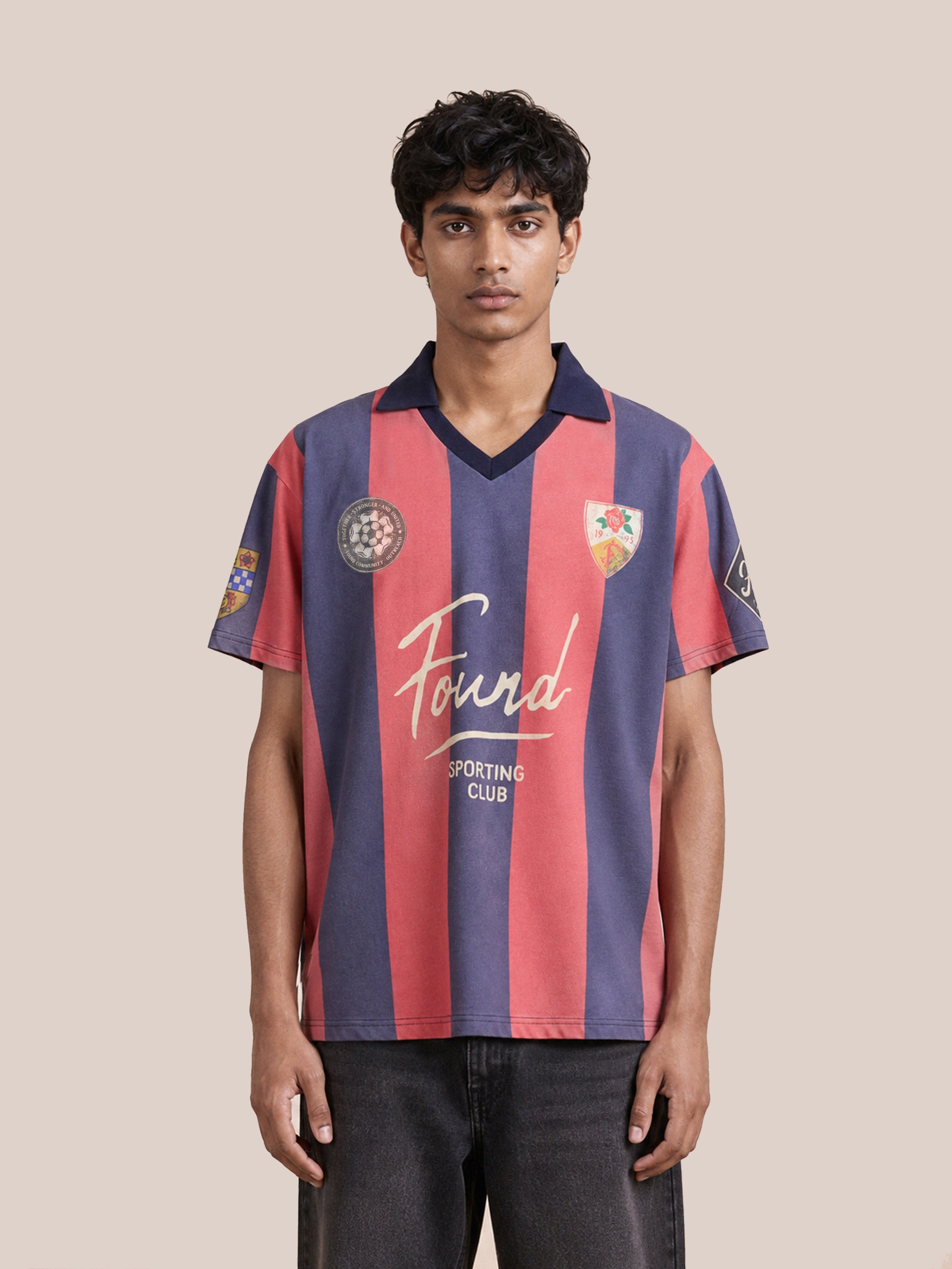 A person stands facing forward against a plain beige background, wearing the FOUND Found Club Short Sleeve Jersey—a red and navy striped football kit adorned with “Found Sporting Club” and various patches on the front.