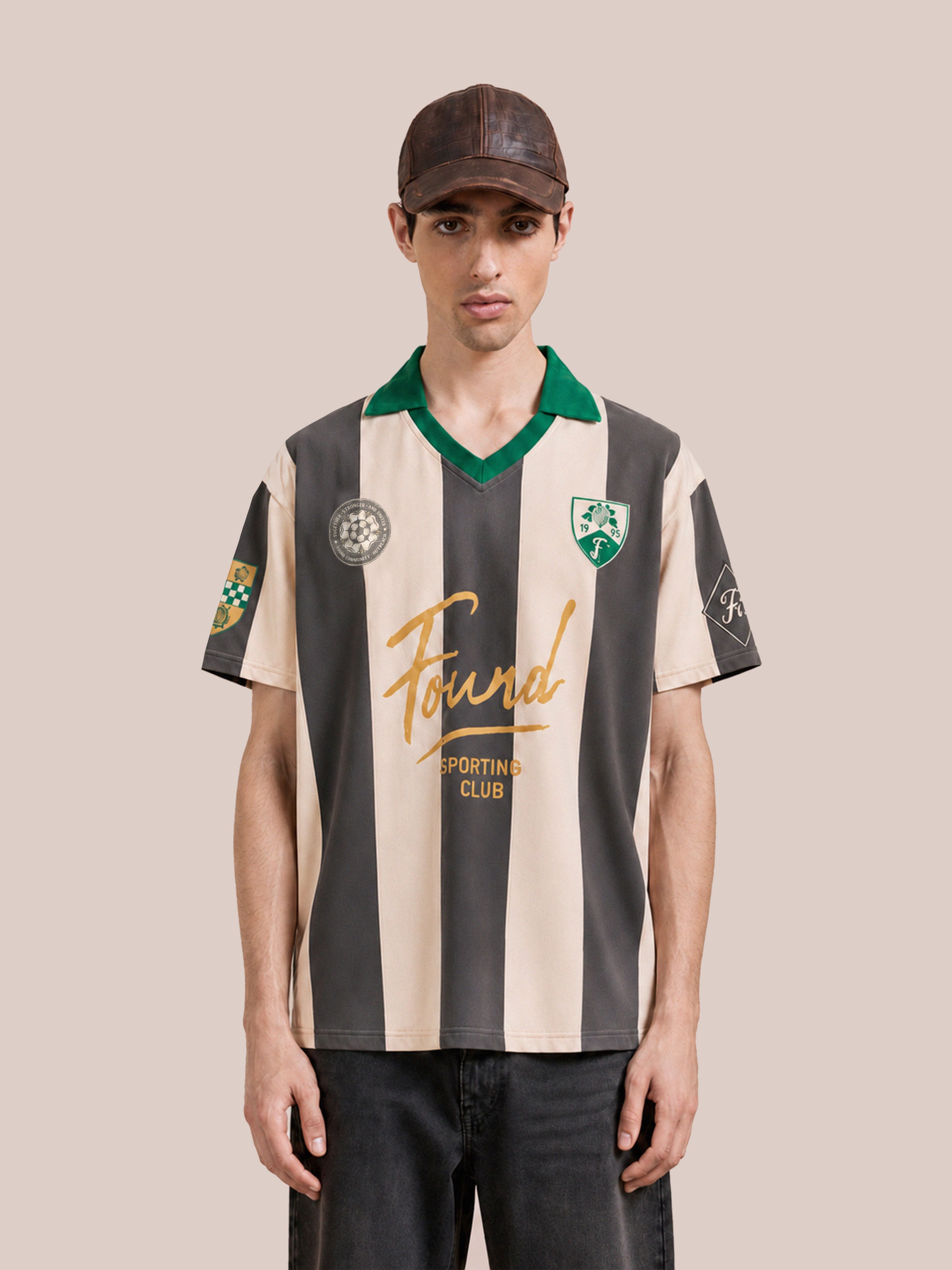 A man wearing the FOUND Found Club Short Sleeve Jersey—a beige and brown striped top with a green collar, multiple logos, and a relaxed unisex fit—stands facing the camera against a plain background, highlighting its modern football-inspired design.