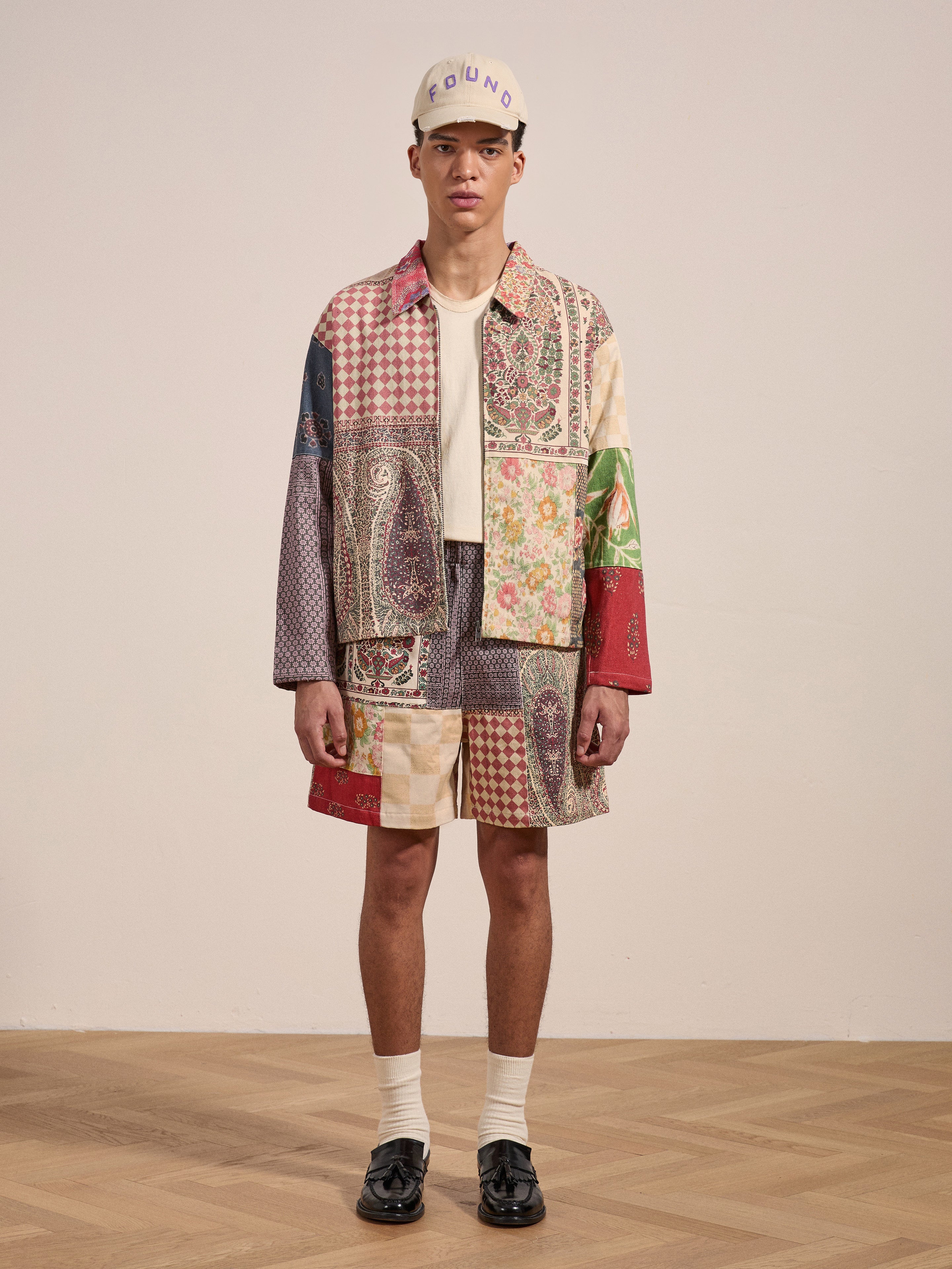 A person stands on a wooden floor wearing a colorful Tapestry Twill Jacket by FOUND and shorts featuring South Asian patterns, paired with a white shirt, black shoes, white socks, and a light-colored cap with text.