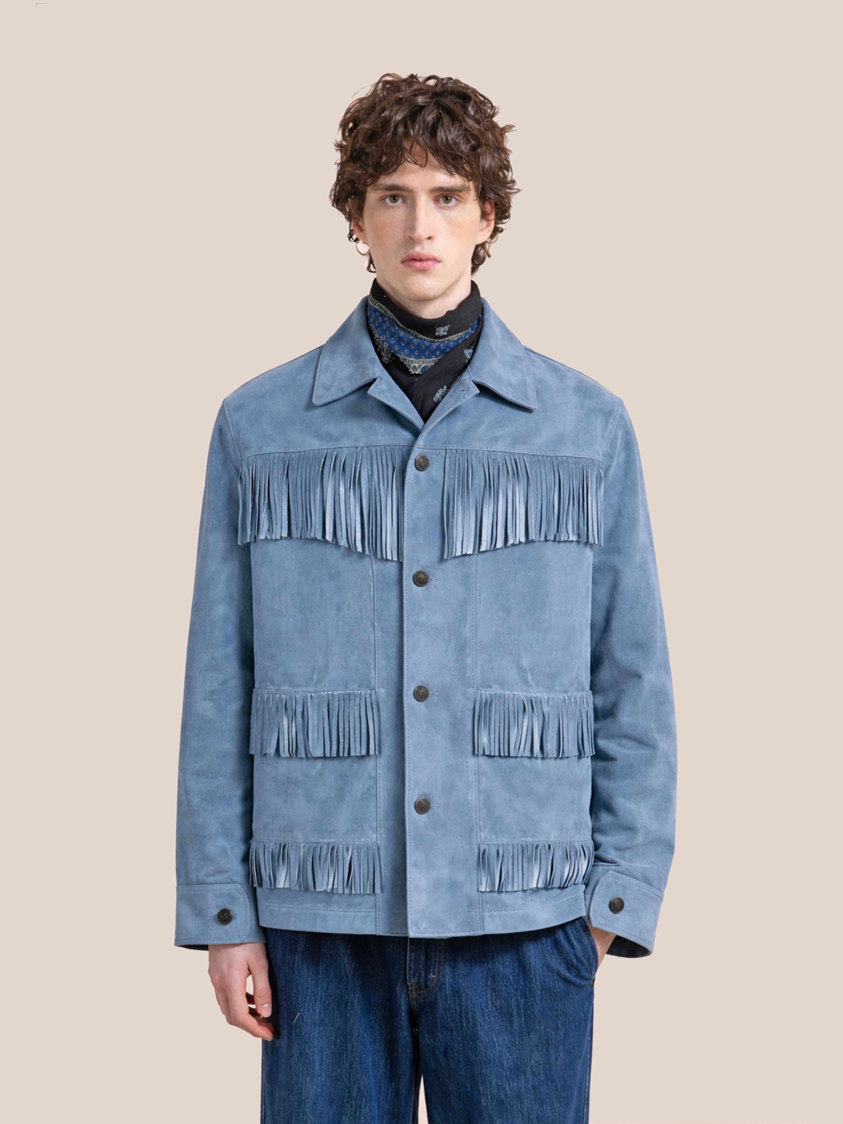 Wearing the FOUND Western Suede Fringe Jacket in indigo blue, styled with a dark neck scarf and blue jeans, this unisex piece features fringe on the chest, pockets, and sleeves—all set against a plain background.