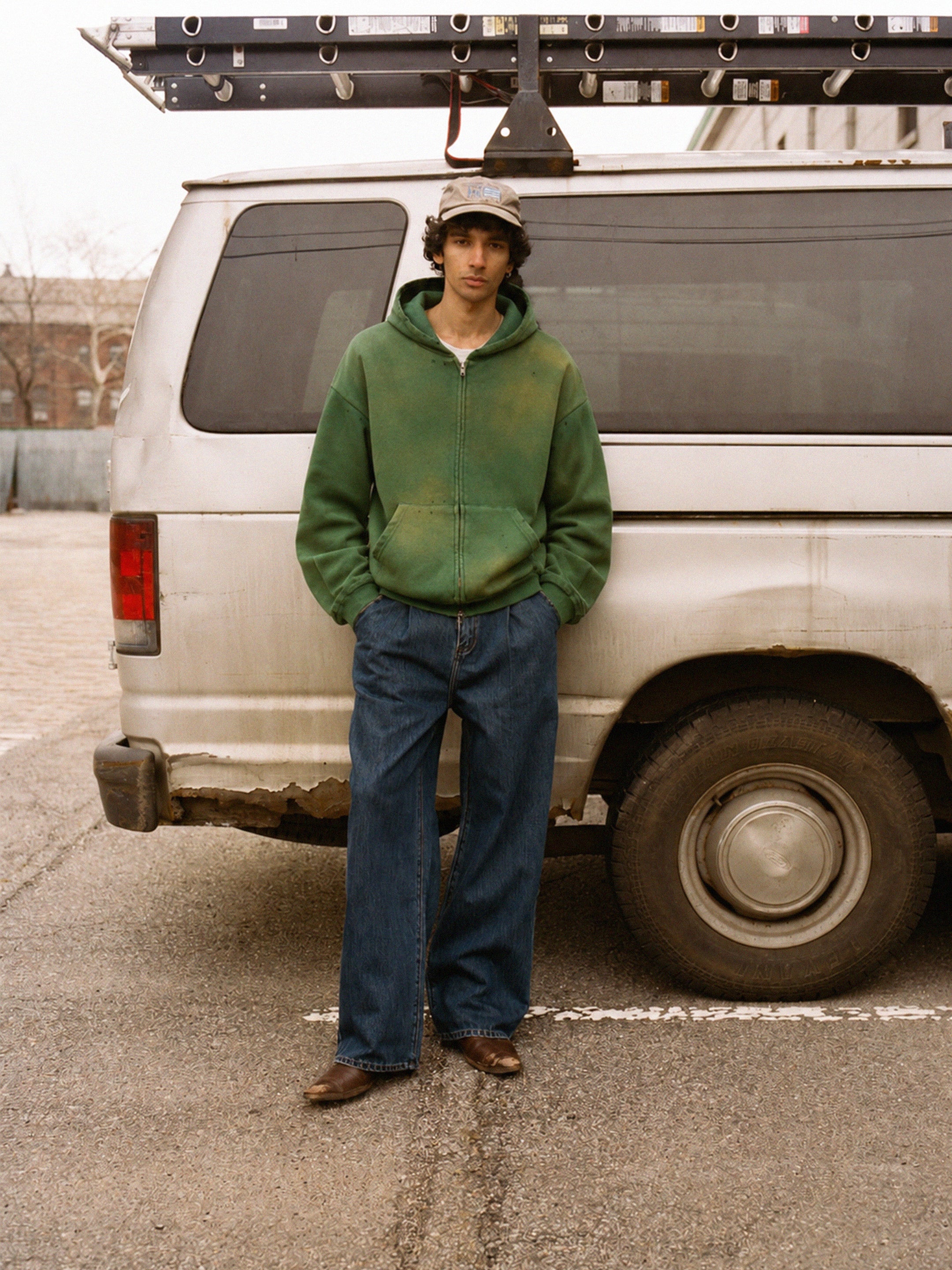 Wearing the FOUND Distressed Thermal Lined Zip Up Hoodie, blue jeans, and brown shoes, a person stands before a white van with a roof ladder in a parking lot.