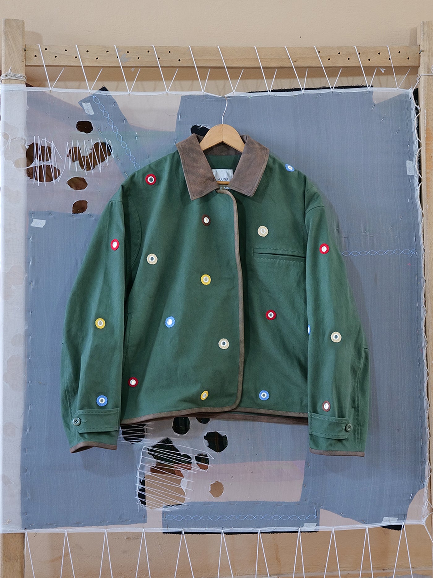 The Hand Embroidered Mirror Abhala Jacket by Found features a brown collar and multicolored circular patches, displayed on a wooden hanger against a textured, stitched fabric background.