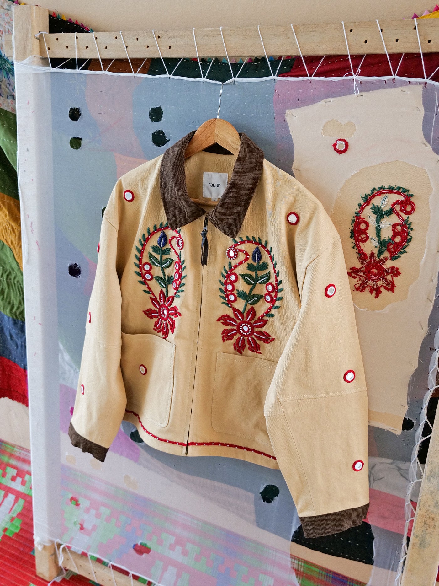 The Found Paisley Motif Hand Embroidered Work Jacket is crafted from beige cotton twill with a brown collar and cuffs, showcasing colorful floral and paisley embroidery, displayed on a wooden hanger against a quilted mesh backdrop.