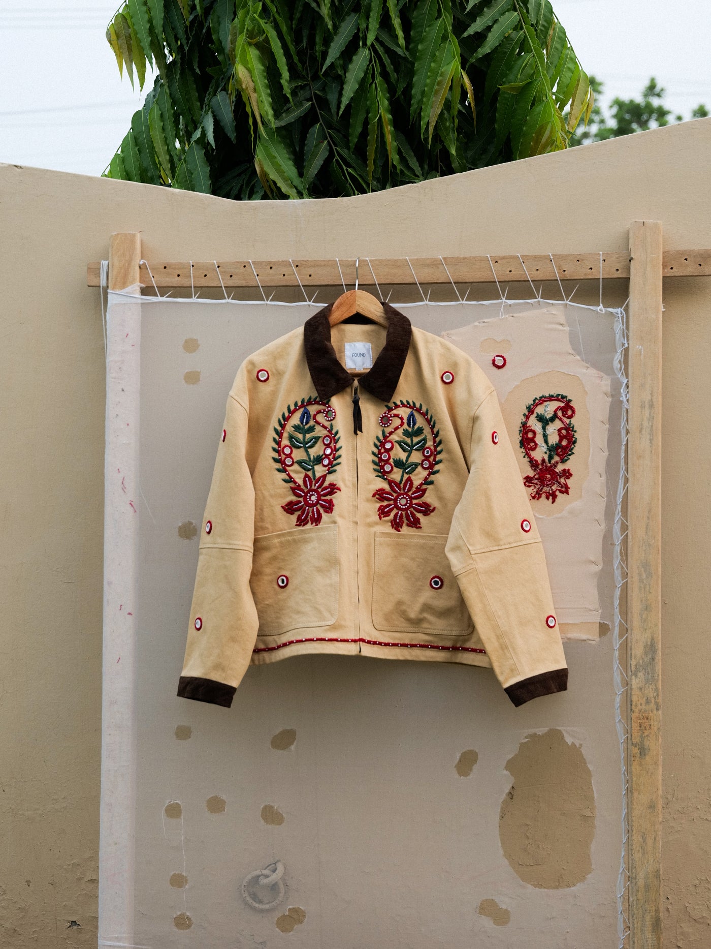 The Found Paisley Motif Hand Embroidered Work Jacket, crafted in beige cotton twill with brown collar and cuffs and red-green paisley embroidery, hangs outdoors on a wooden frame against a beige wall.
