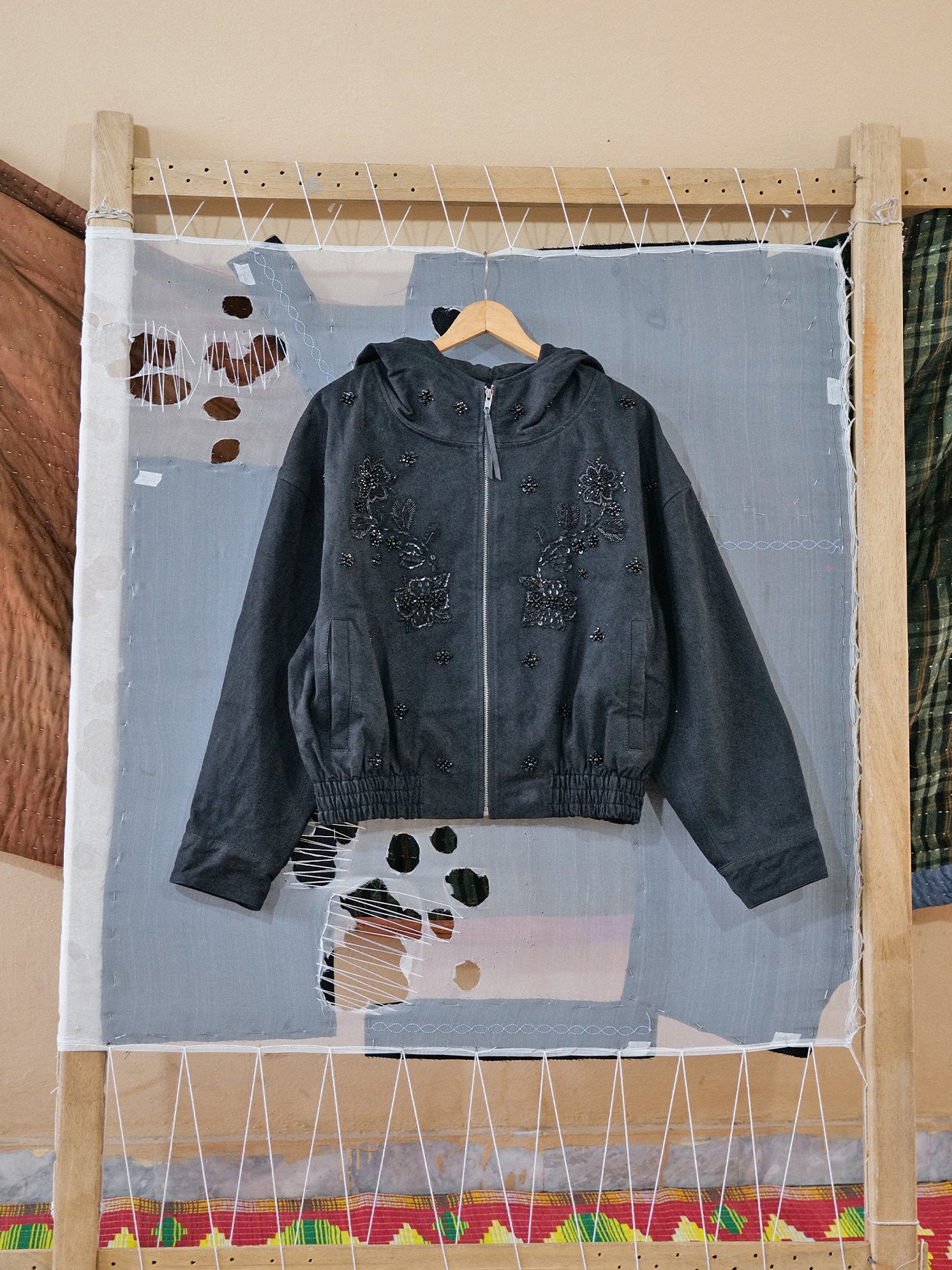 The FOUND Hand Embroidered Tonal Anorak Jacket, a dark gray unisex cotton piece, hangs on a wooden hanger against a mixed-media textile backdrop.