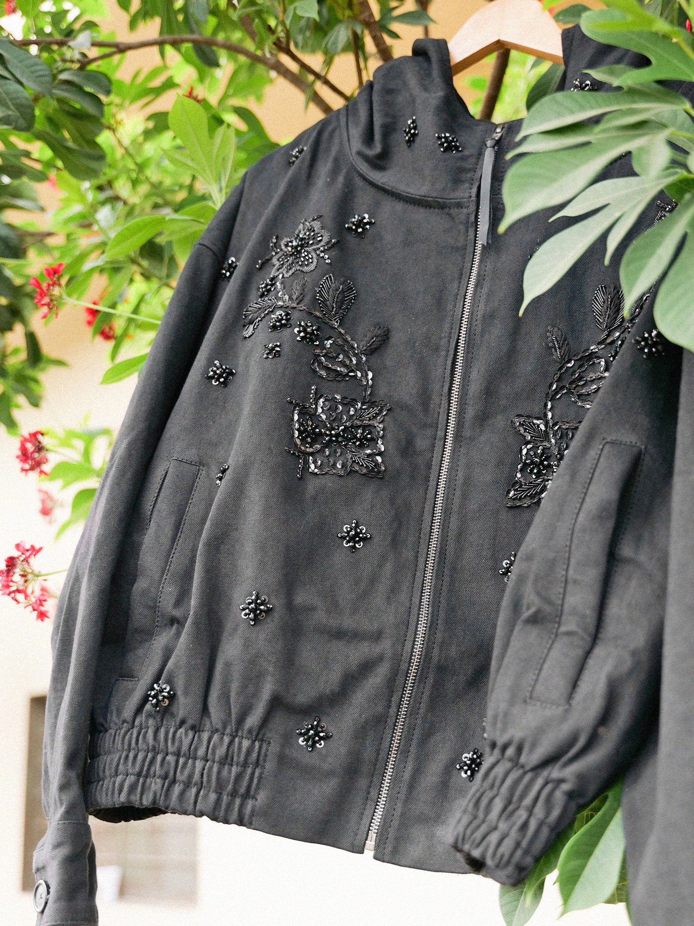 The FOUND Hand Embroidered Tonal Anorak Jacket, a black unisex cotton piece with Salma Sitara embroidery and beaded florals, hangs on a wooden hanger amid green leaves and small red flowers.