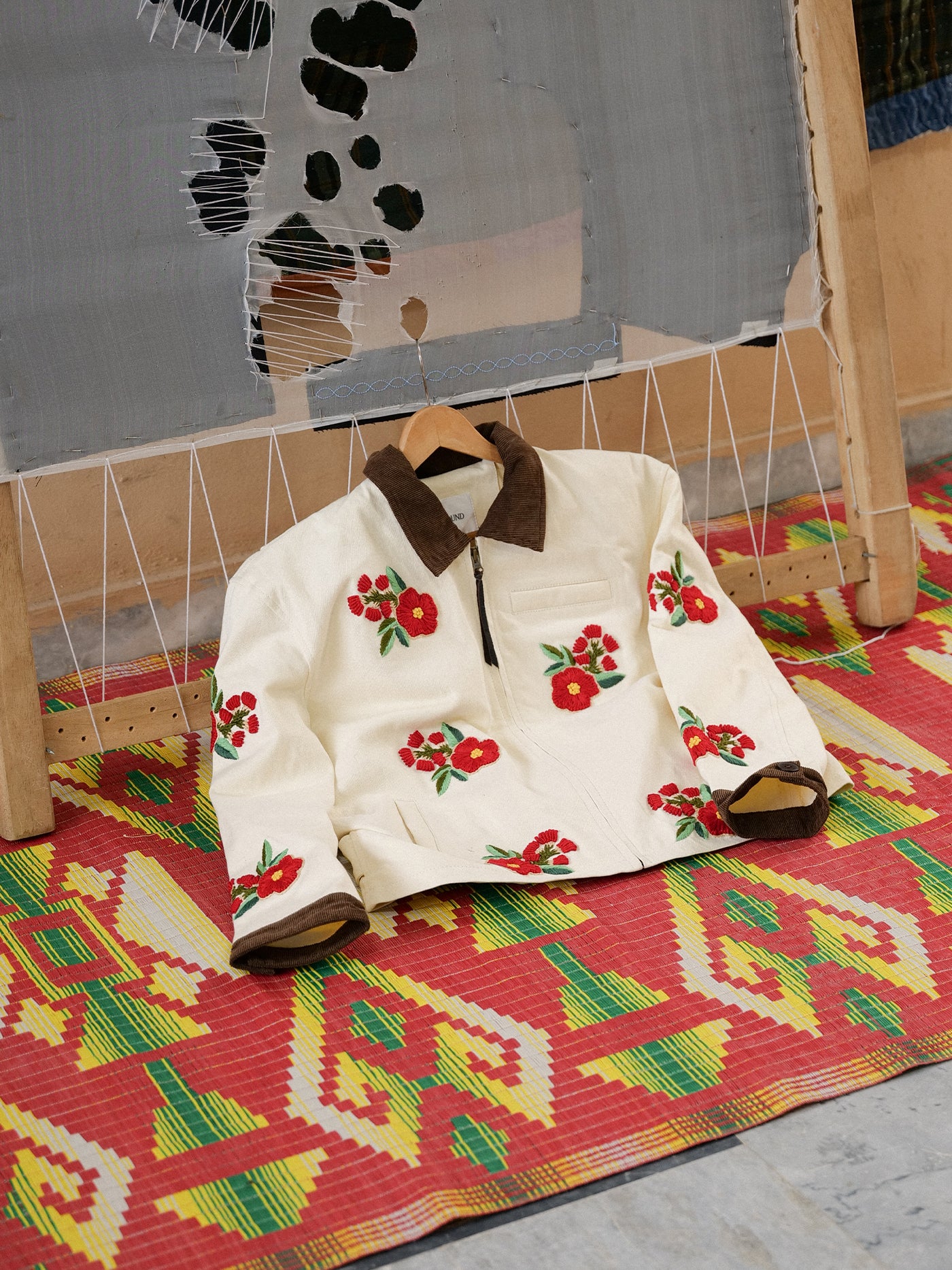 The Floral Hand Embroidered Work Jacket by Found, cream-colored with red floral motifs and brown collar and cuffs, handmade in Pakistan, hangs on a wooden hanger atop a patterned mat before textile art.