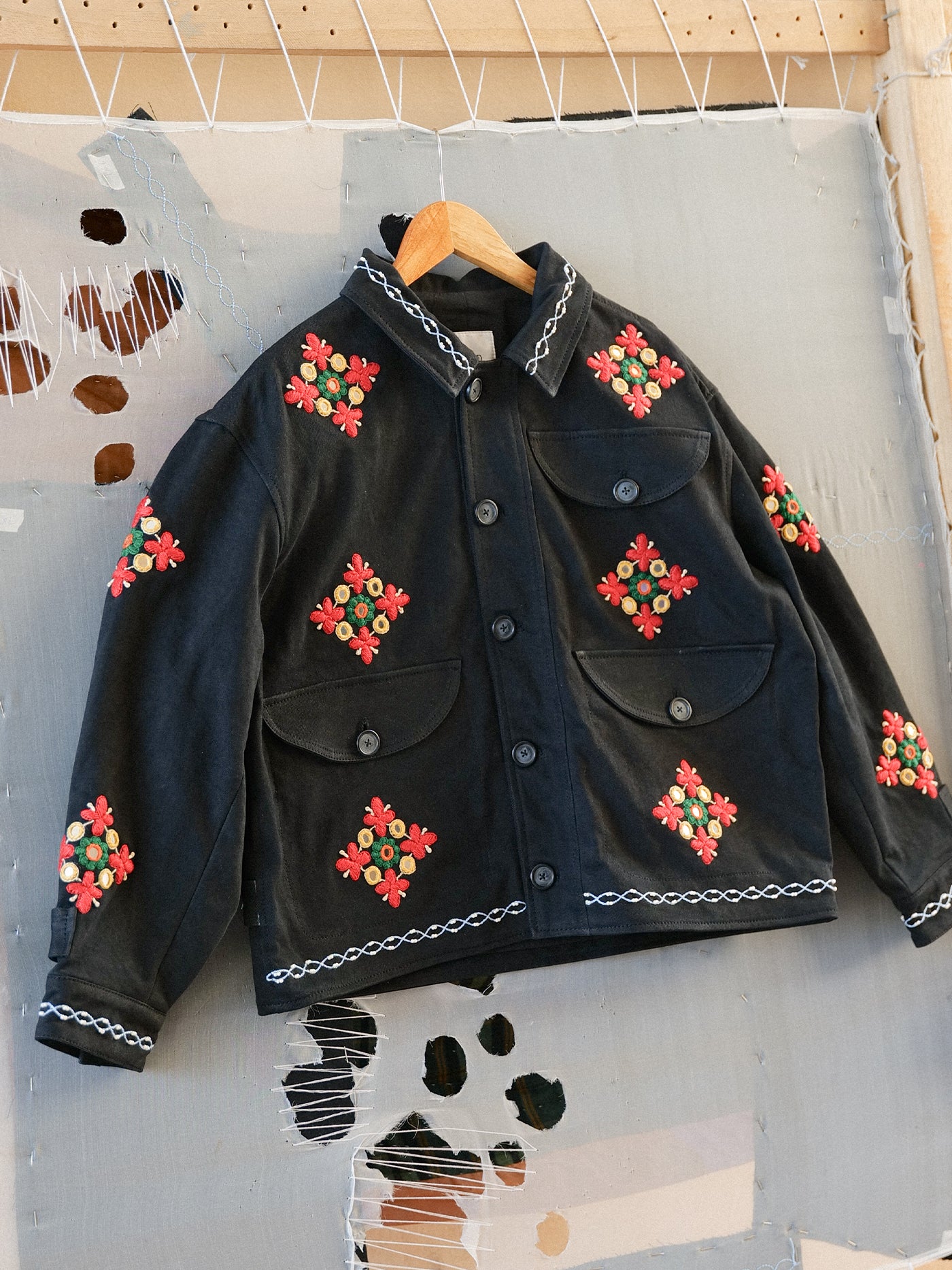 The Found Mirror Ornament Hand Embroidered Work Jacket, crafted from black cotton twill with red and green floral embroidery, hangs on a wooden hanger against a gray textured background.