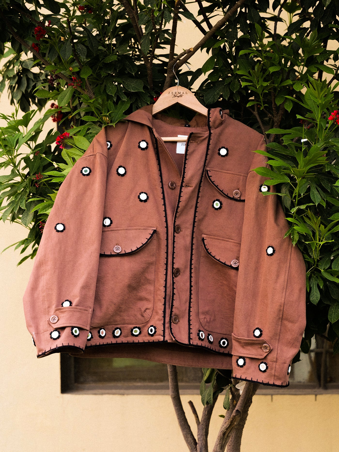 The FOUND Hand Embroidered Hooded Mirror Anorak, featuring large front pockets and white mirror flower embroidery inspired by South Asian craft, hangs on a wooden hanger against leafy green shrubbery.