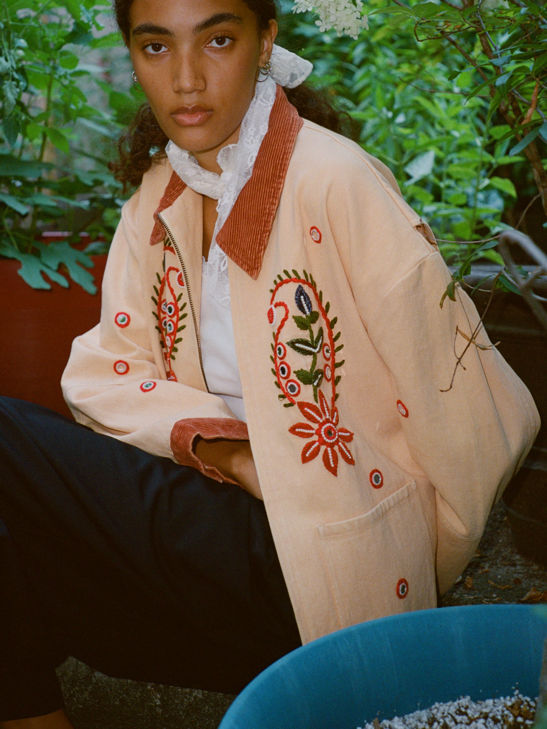 A person sits outdoors among potted plants wearing the Found Paisley Motif Hand Embroidered Work Jacket, which features red and green patterns with artisan detail, a white lace collar, and dark pants.