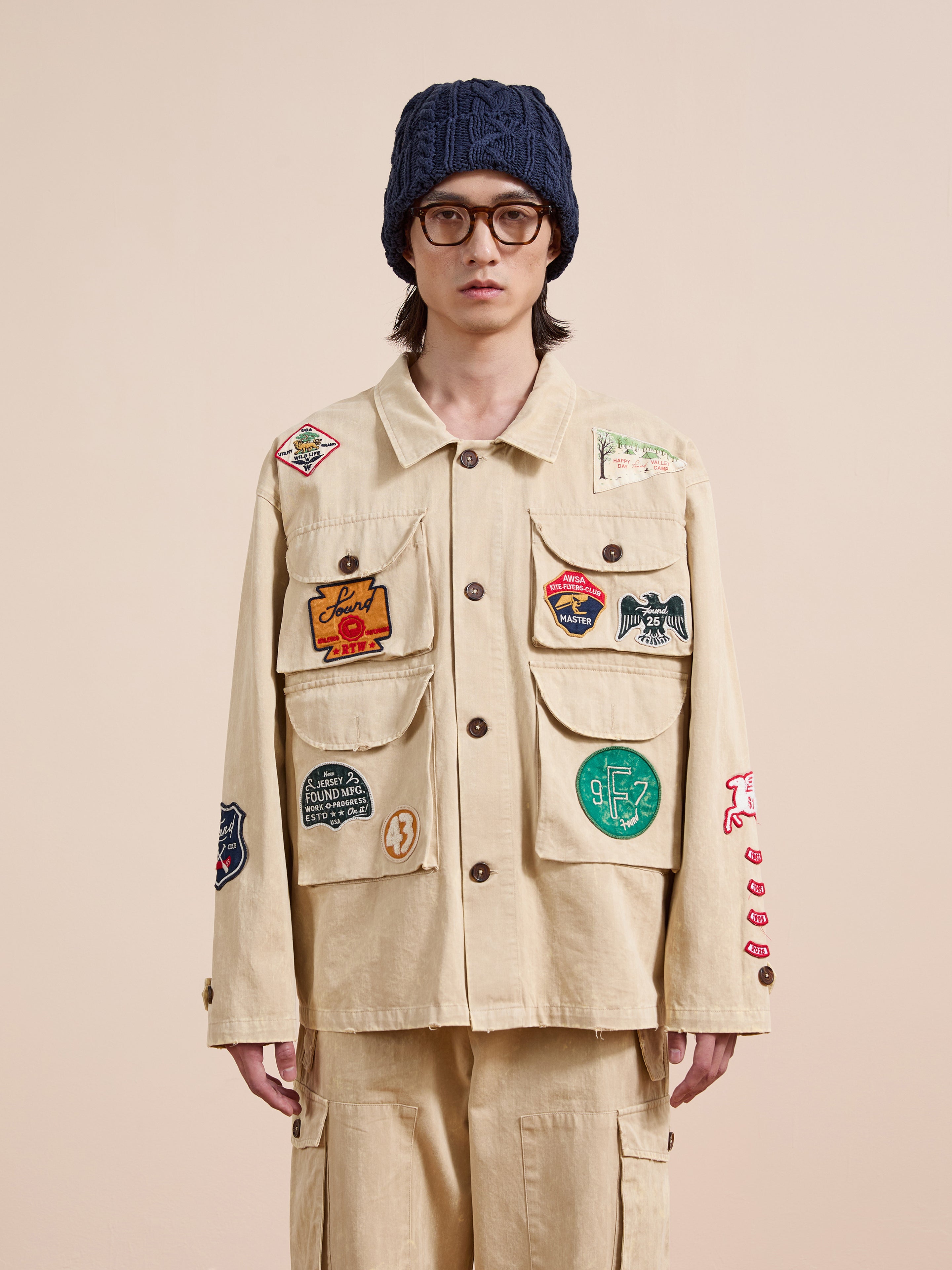 A person wearing the FOUND Herringbone Utility Patchwork Jacket, matching pants, a navy knit beanie, and round glasses stands against a plain background.