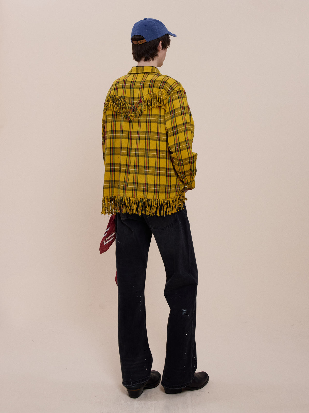 Person facing away in a yellow plaid FOUND High Plains Fringe Shirt, black jeans, black shoes, blue cap, and red bandana in pocket against a plain background.
