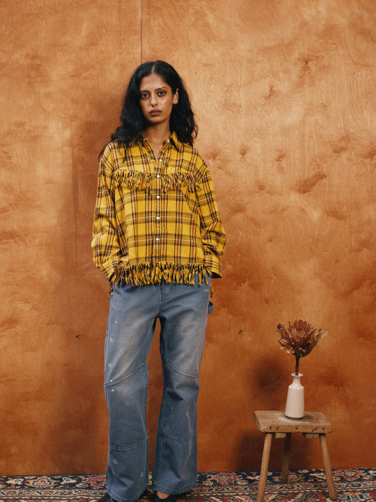 Woman wearing the FOUND High Plains Fringe Shirt and blue jeans stands against a wooden backdrop next to a stool with a vase and dried flower.
